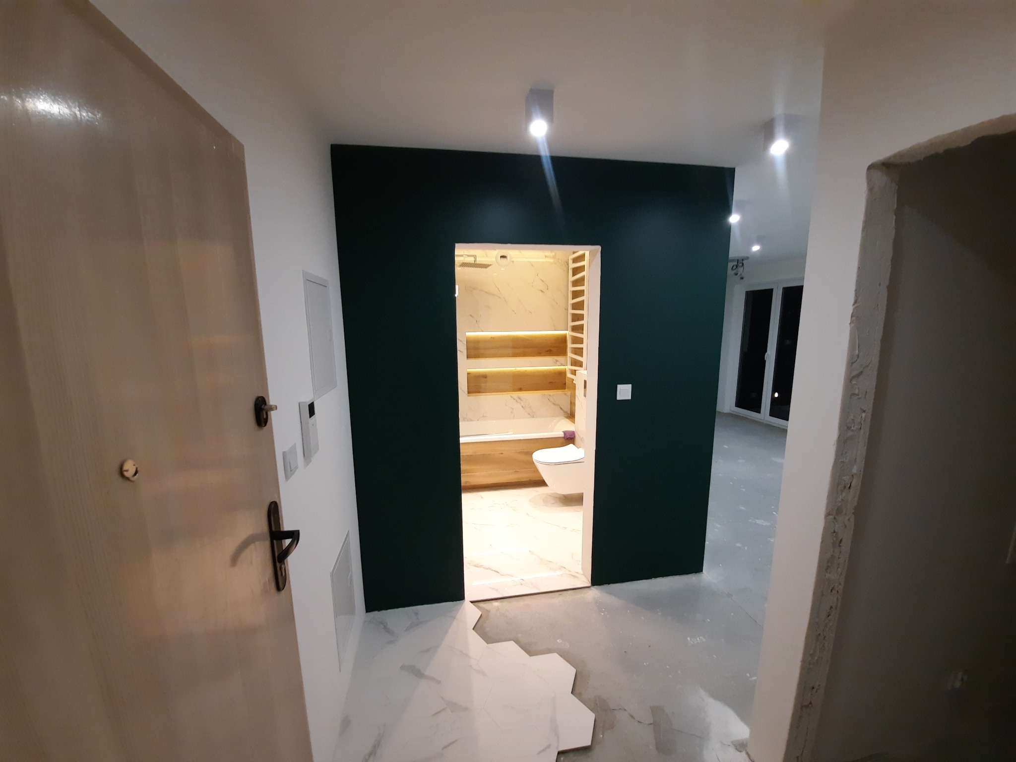 Widok na wykończoną łazienkę z marmurowymi płytkami, podświetlanymi półkami i toaletą, przez ciemnozielone obramowanie wejścia, z fragmentem otwartego pokoju i kładzioną podłogą w tle.