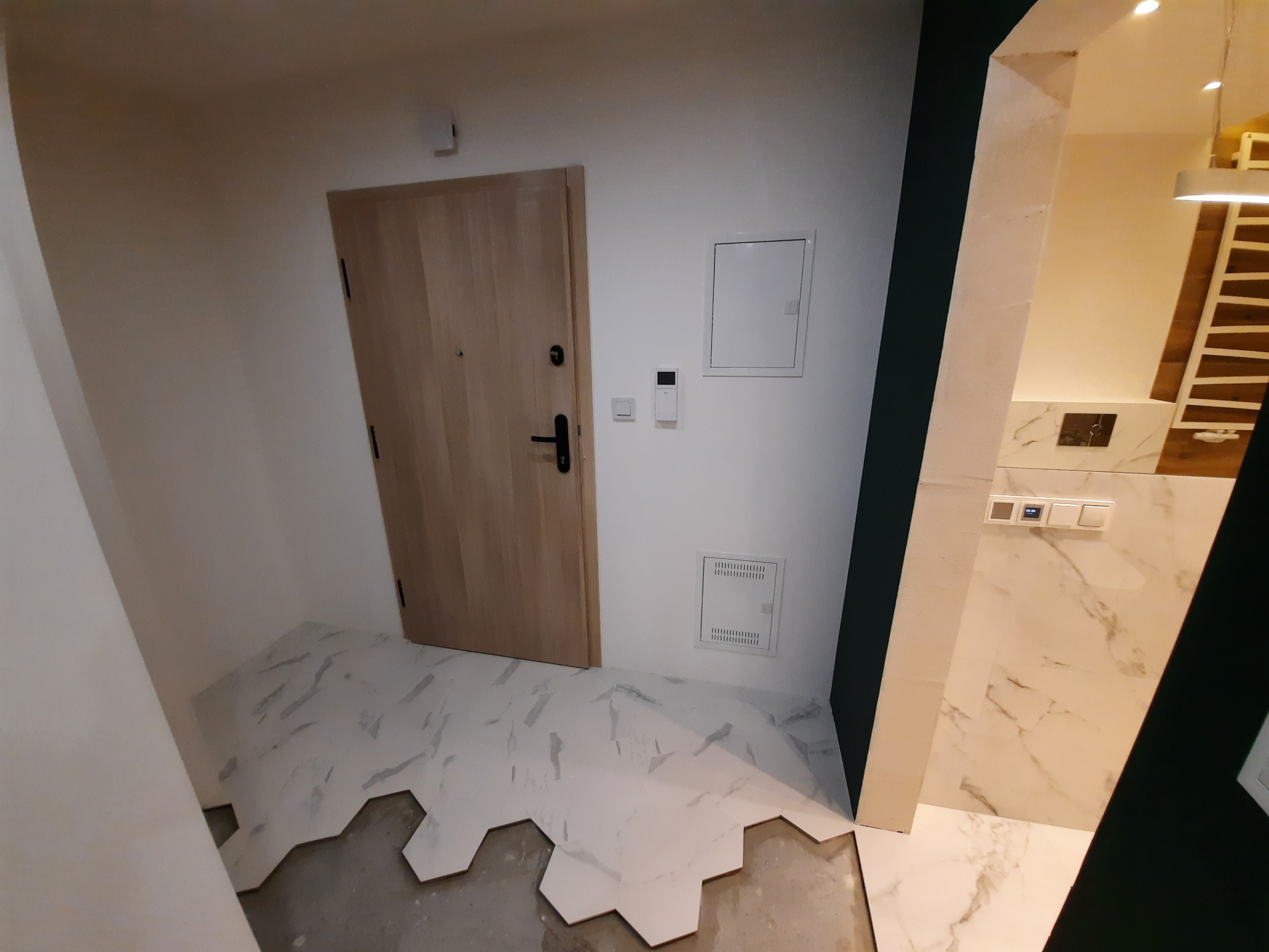 Wnętrze w trakcie wykańczania: ułożona częściowo podłoga z heksagonalnych płytek imitujących marmur, widoczna drewniana ościeżnica drzwiowa oraz fragment marmurowej ściany z włącznikami światła.