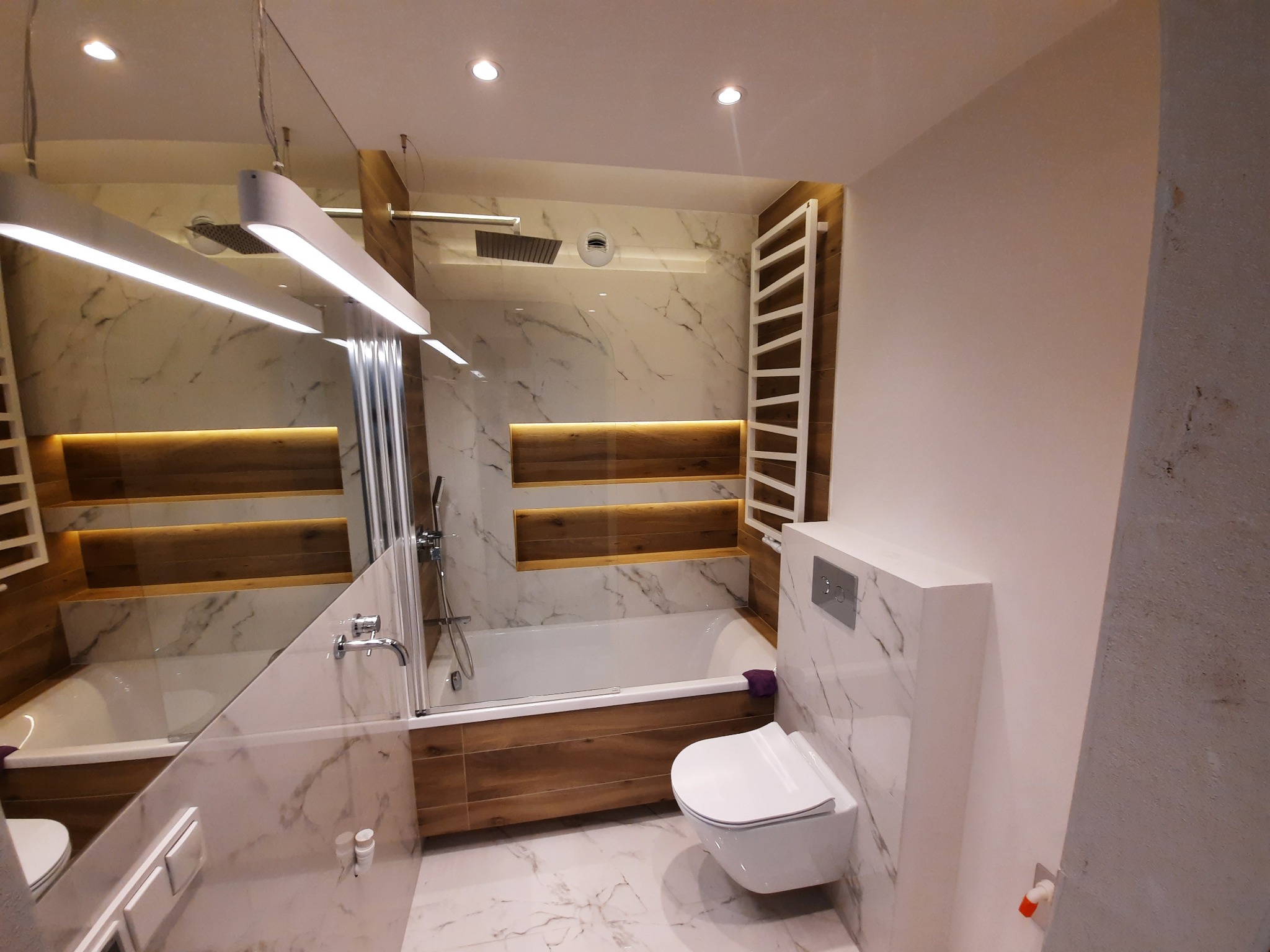 Wykończona łazienka z wanną obudowaną drewnopodobnymi panelami, marmurowymi płytkami na ścianach i podłodze, podwieszaną toaletą i dekoracyjnym oświetleniem LED za wanną.