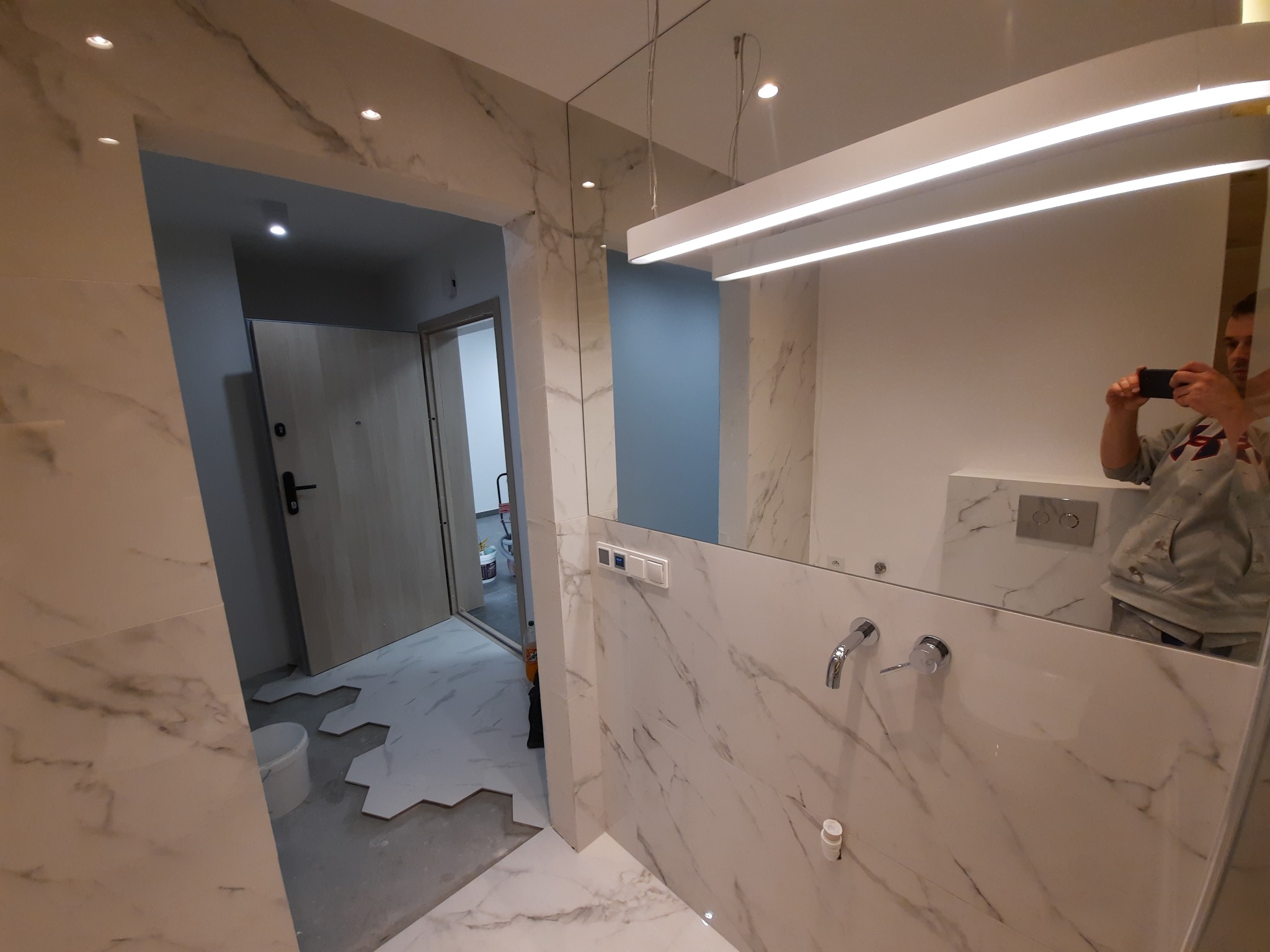 Łazienka w trakcie remontu w Tworkowej: widoczne białe marmurowe płytki na ścianach, nowoczesna armatura, lustro z oświetleniem LED oraz fragment podłogi z ułożonymi sześciokątnymi płytkami, w tle...