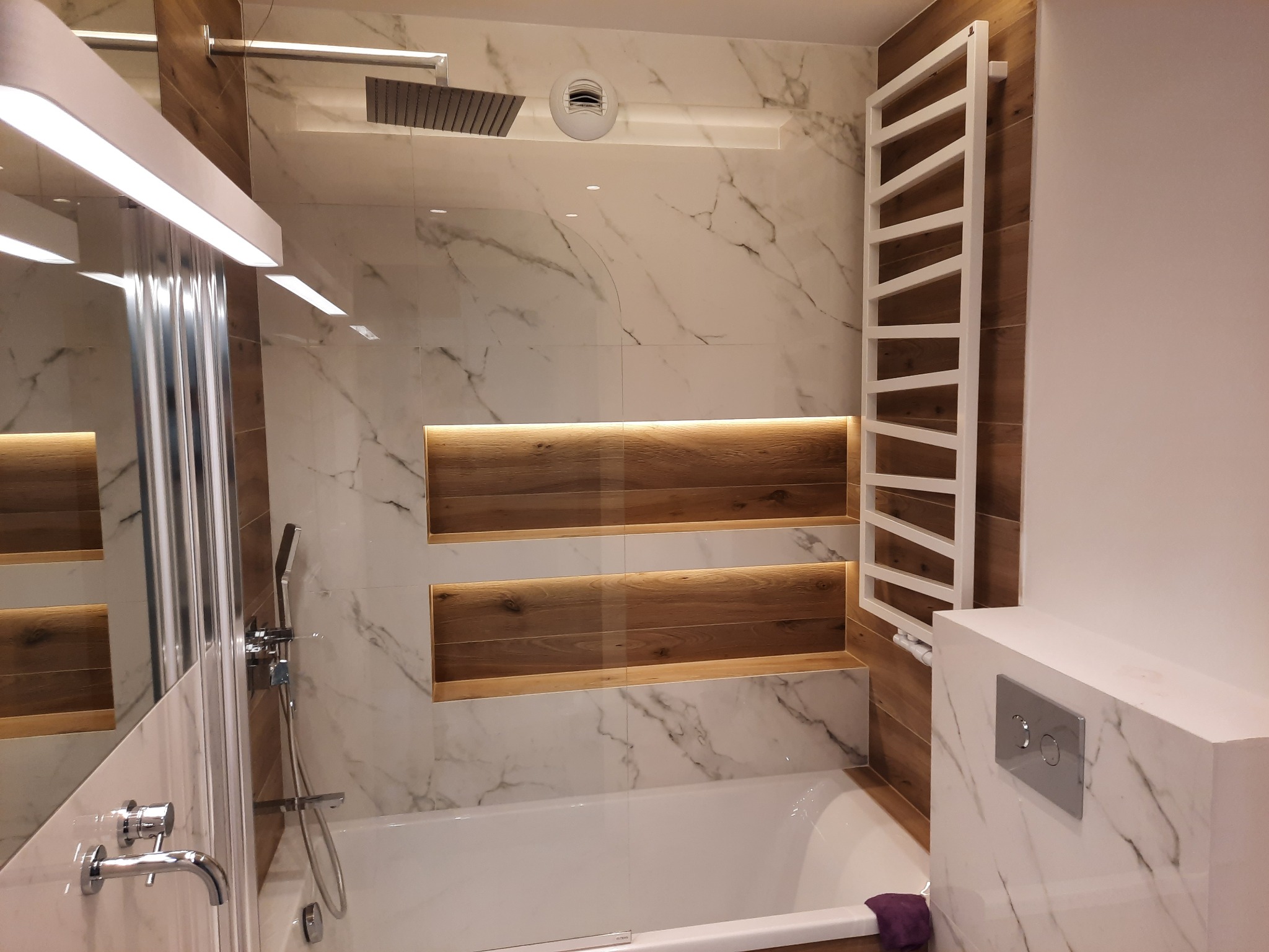 Nowoczesna łazienka z wanną, marmurowymi płytkami, drewnianymi wnękami z oświetleniem LED, grzejnikiem drabinkowym i deszczownicą.
