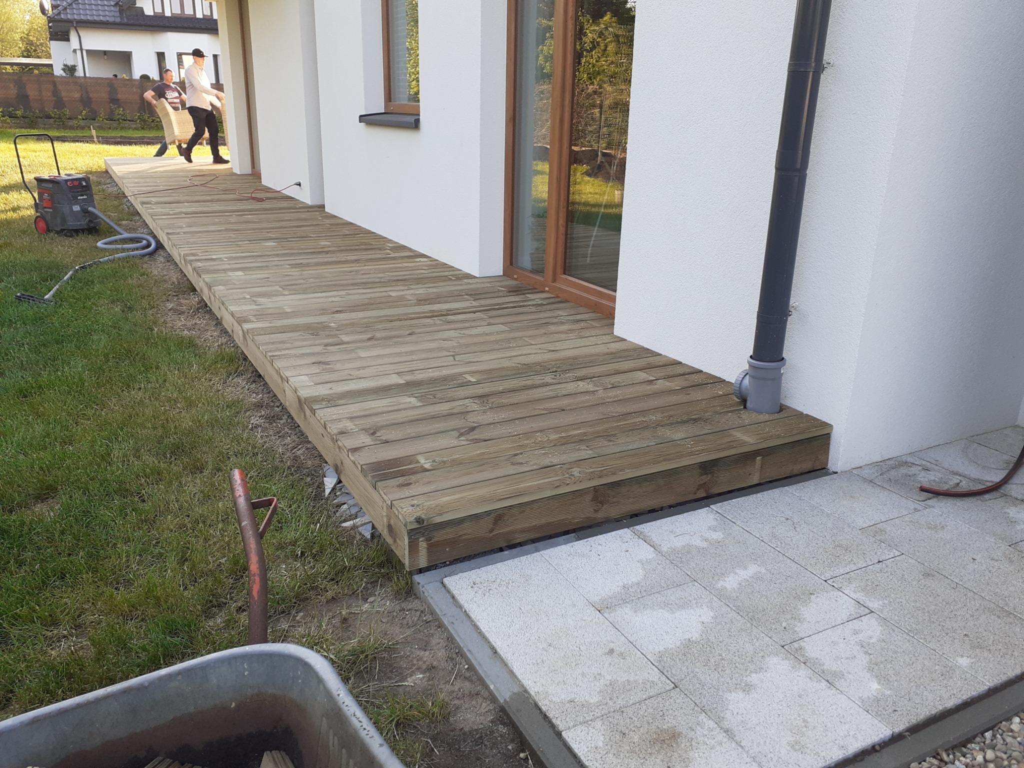 Nowo wybudowany, drewniany taras przy białym domu, z widocznym odkurzaczem przemysłowym i taczką; w tle dwie osoby przenoszą meble.