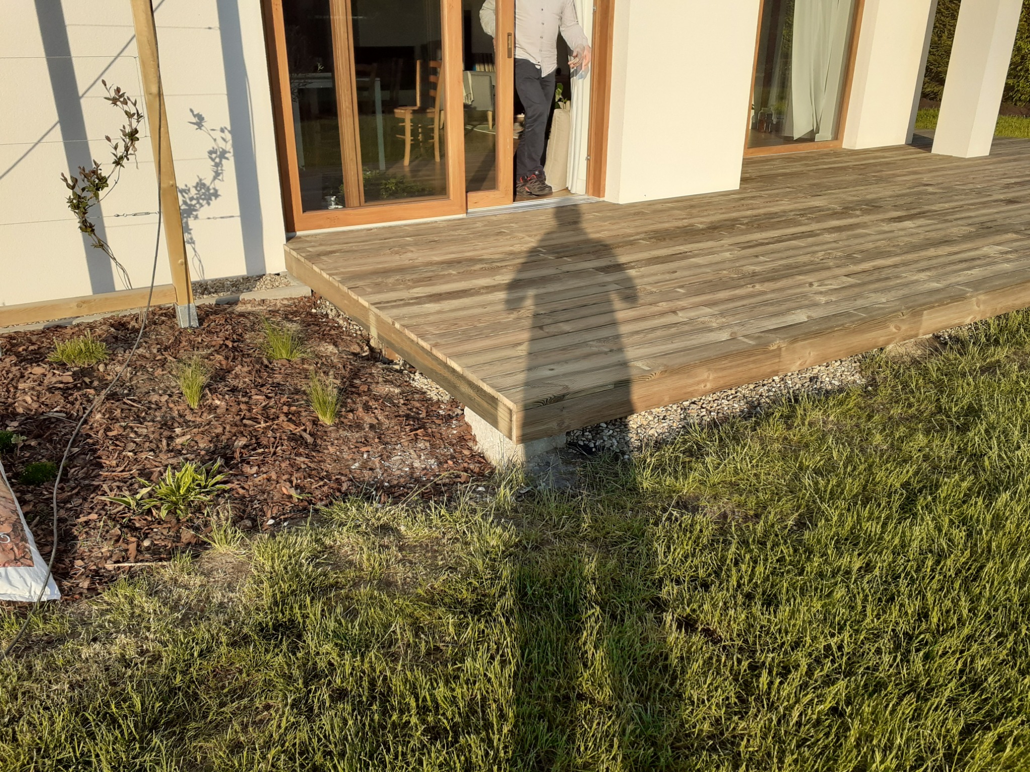Nowo wybudowany drewniany taras przy domu z przesuwnymi drzwiami balkonowymi, widoczny cień osoby stojącej w drzwiach, świeżo posadzone rośliny w korze.