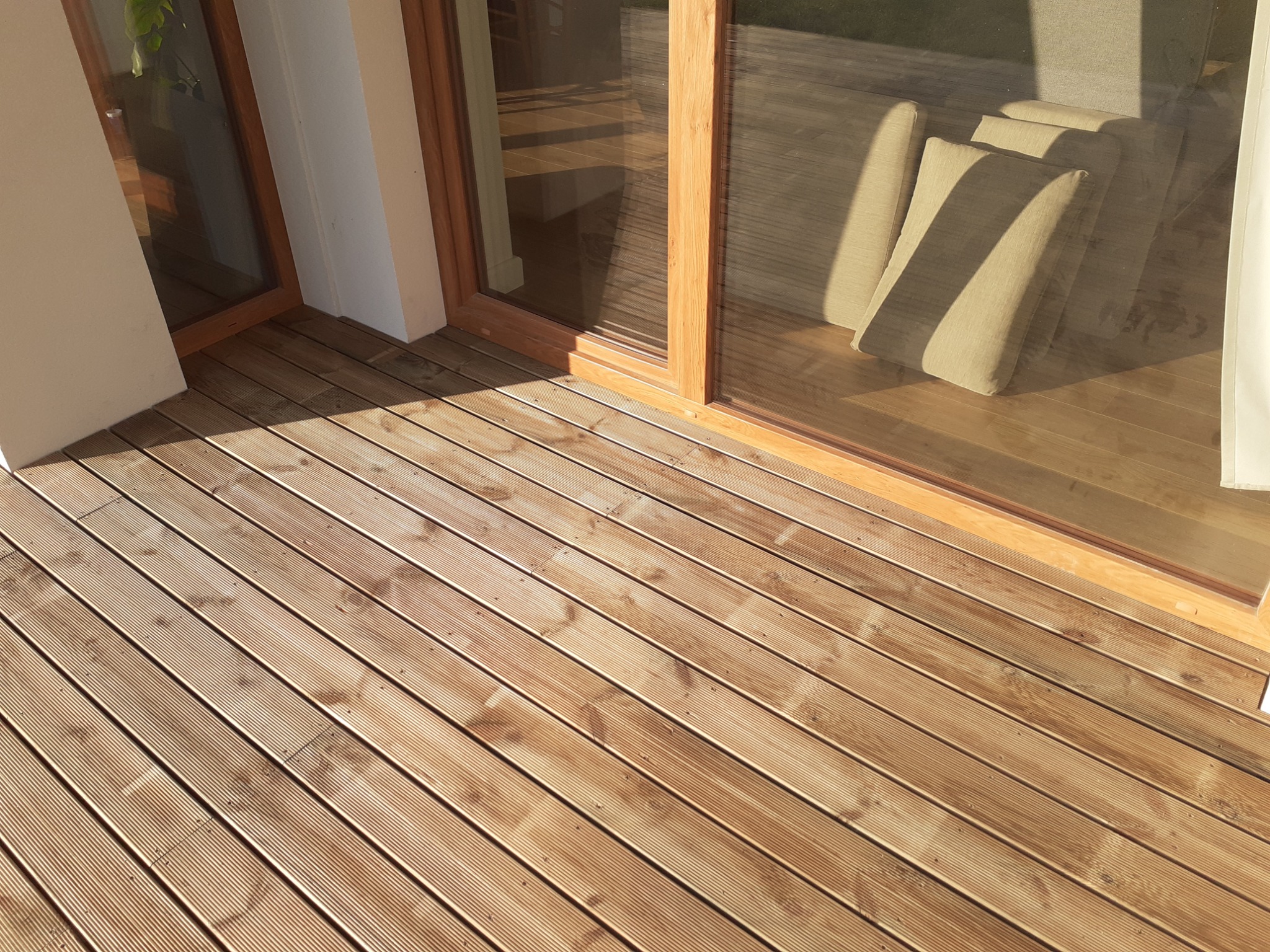 Nowoczesny taras z drewnianą podłogą z desek, widok na przesuwne drzwi tarasowe z drewnianą ramą i jasnymi poduszkami wewnątrz.
