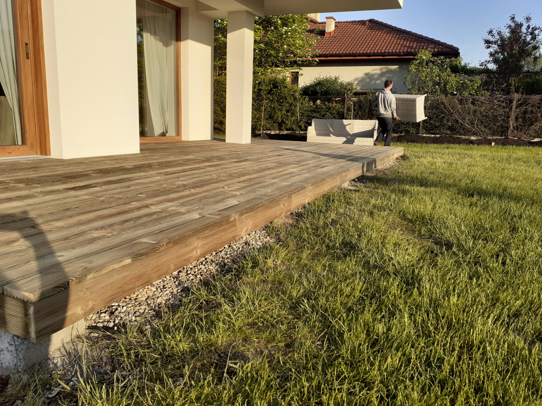 Drewniany taras przydomowy z widoczną konstrukcją, obok trawnik i osoba przenosząca meble ogrodowe w tle.