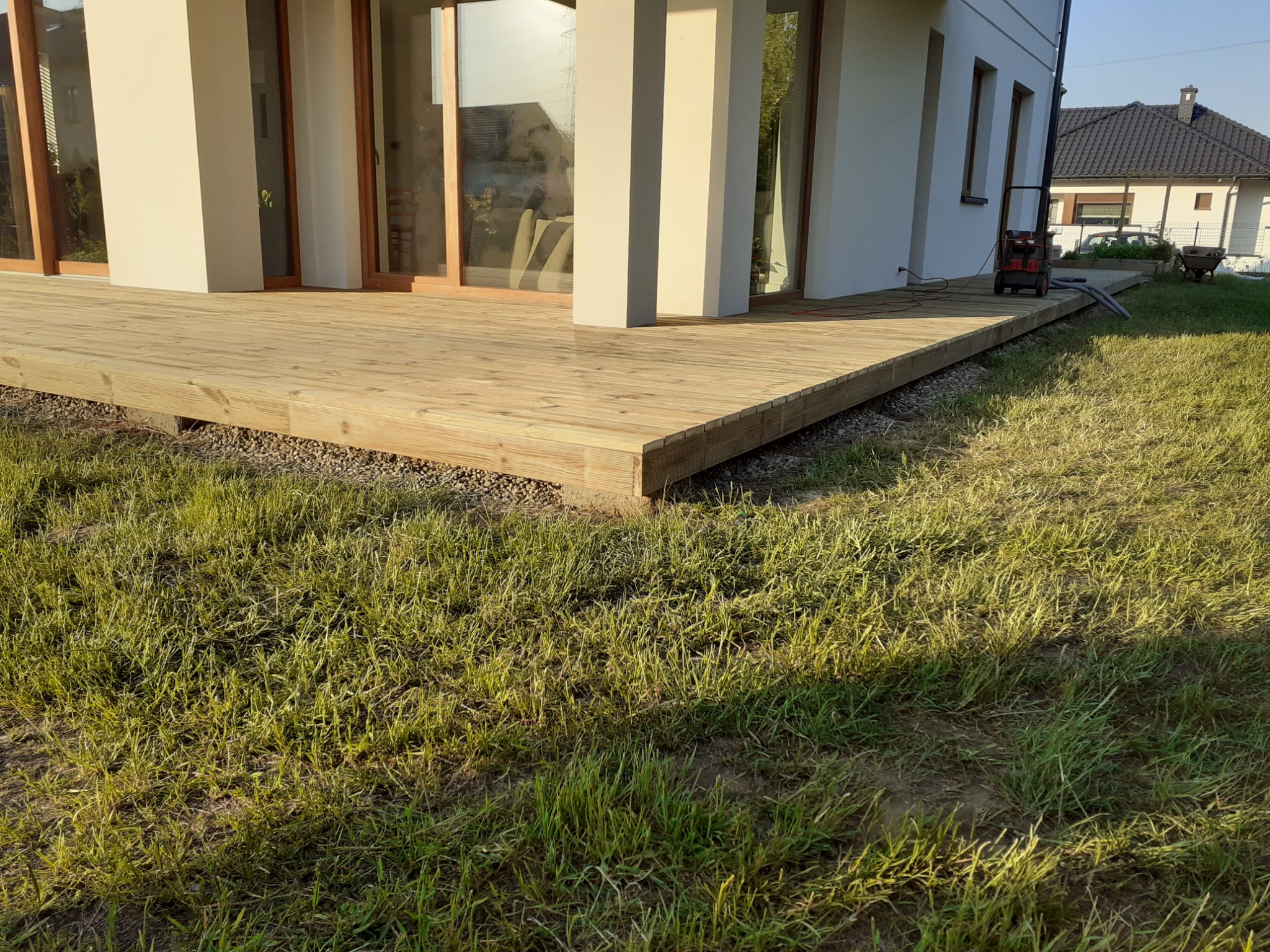 Nowoczesny taras z jasnego drewna przylegający do białego domu z dużymi oknami przesuwnymi, widoczna kosiarka elektryczna na tarasie i trawnik.