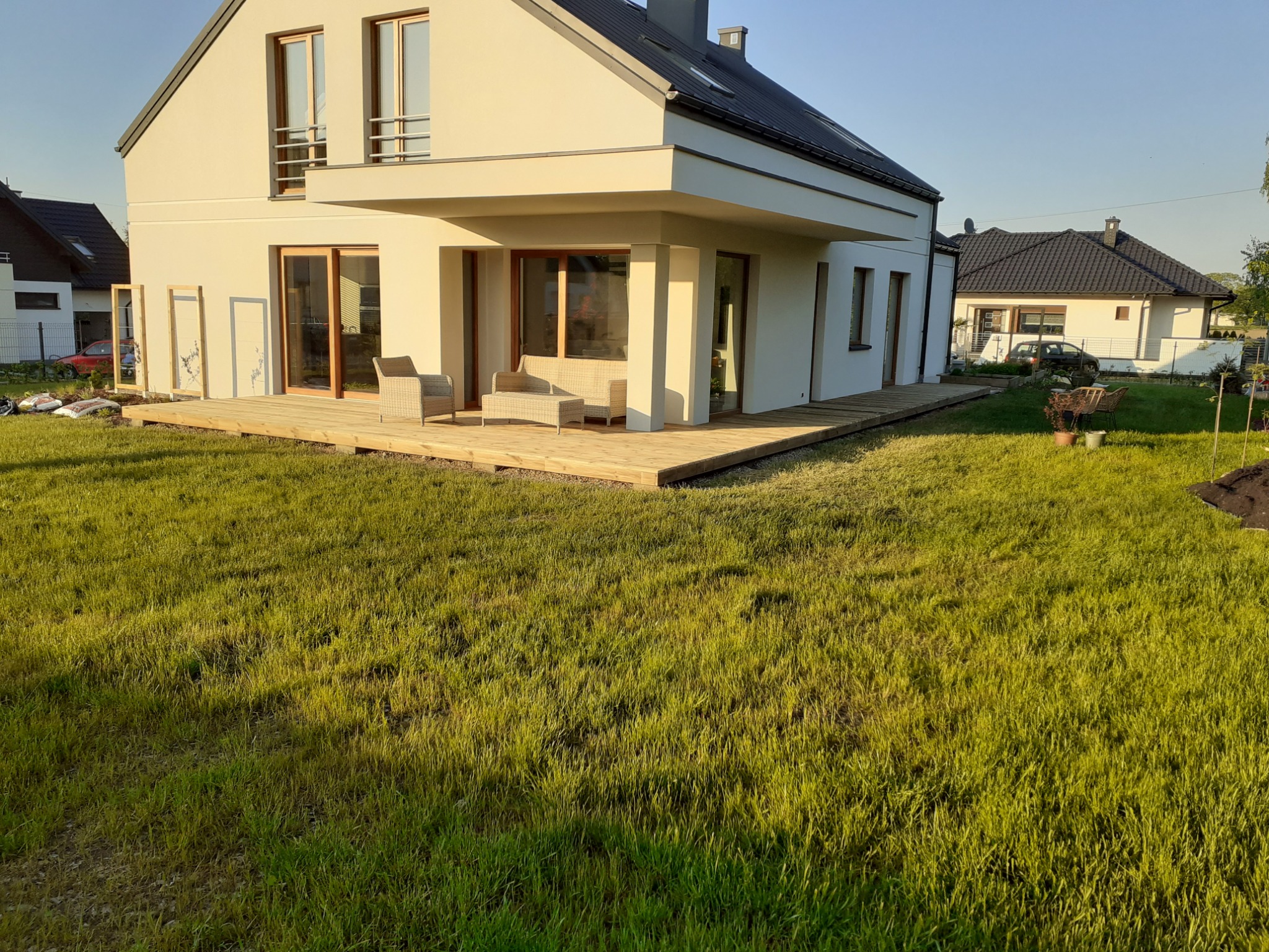 Nowoczesny dom z jasną elewacją i drewnianym tarasem, na którym znajdują się rattanowe meble ogrodowe, widok z trawnika w słoneczny dzień.