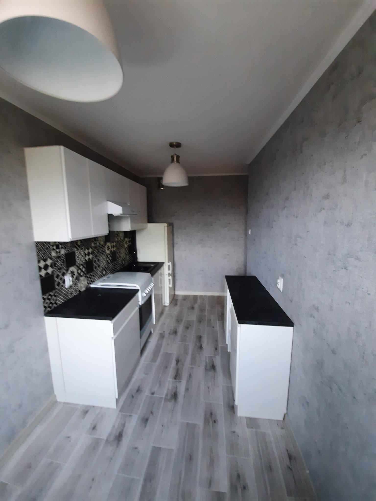 Wnętrze kuchni po remoncie, widok na białe szafki z czarnymi blatami, wzorzyste płytki nad blatem, szare ściany z efektem betonu i podłogę imitującą drewno.