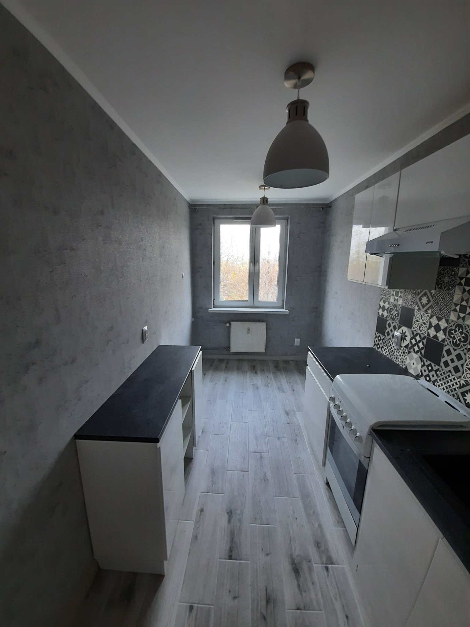 Wykończona kuchnia z szarą, teksturowaną ścianą, białymi szafkami z czarnymi blatami, wzorzystymi płytkami nad kuchenką i dwoma wiszącymi lampami w stylu industrialnym.