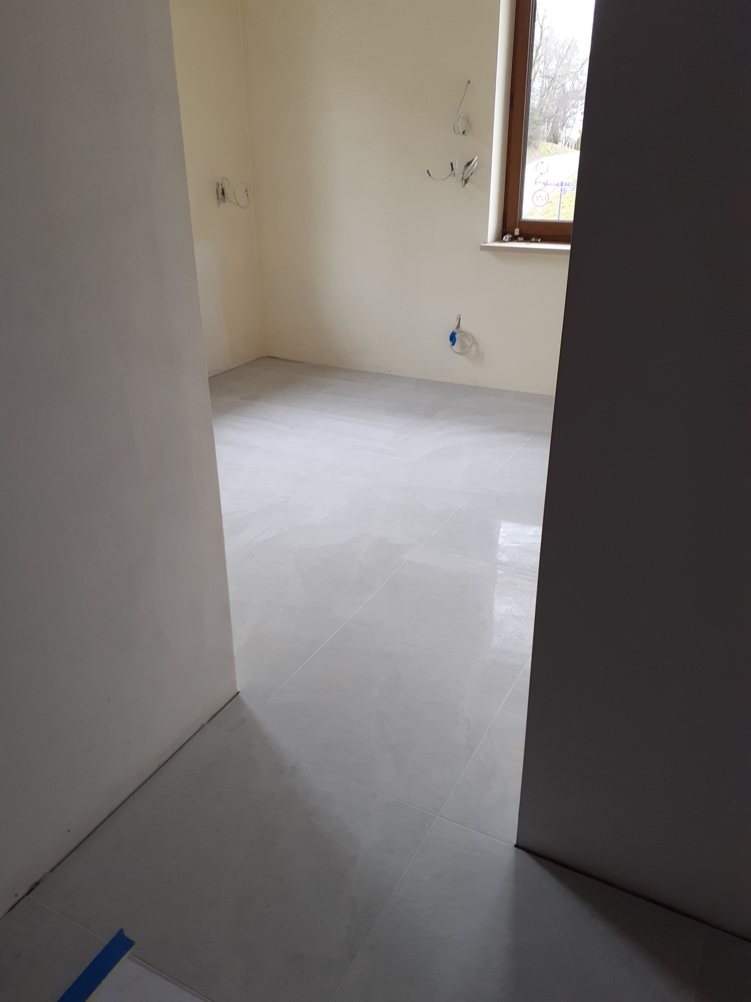 Jasne, błyszczące płytki podłogowe ułożone w nowym pomieszczeniu, widoczne przewody elektryczne wystające ze ściany, okno z widokiem na drzewa.