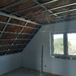 Montaż płyt karton gips na poddaszu: widoczna konstrukcja metalowa, ocieplenie wełną mineralną, przewody elektryczne i okno z widokiem na zewnątrz.