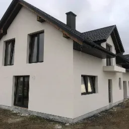 Nowoczesny dom jednorodzinny z jasną elewacją o ziarnistej fakturze, ciemną dachówką i czarnymi oknami, widok z boku na ukończony budynek.