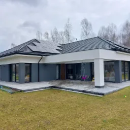 Nowoczesny, parterowy dom z szarymi ścianami, panelami słonecznymi na dachu i tarasem wyłożonym kostką, otoczony trawnikiem i drzewami.