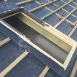 Montaż okna dachowego: widok na konstrukcję dachu z membraną, drewnianą ramą okienną i łatami. W tle widoczne narzędzia i akcesoria montażowe.