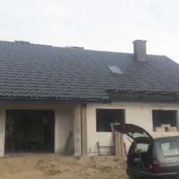 Nowy, grafitowy dach z oknem połaciowym na surowym domu jednorodzinnym w trakcie budowy, widok z boku z otwartą klapą bagażnika samochodu osobowego.