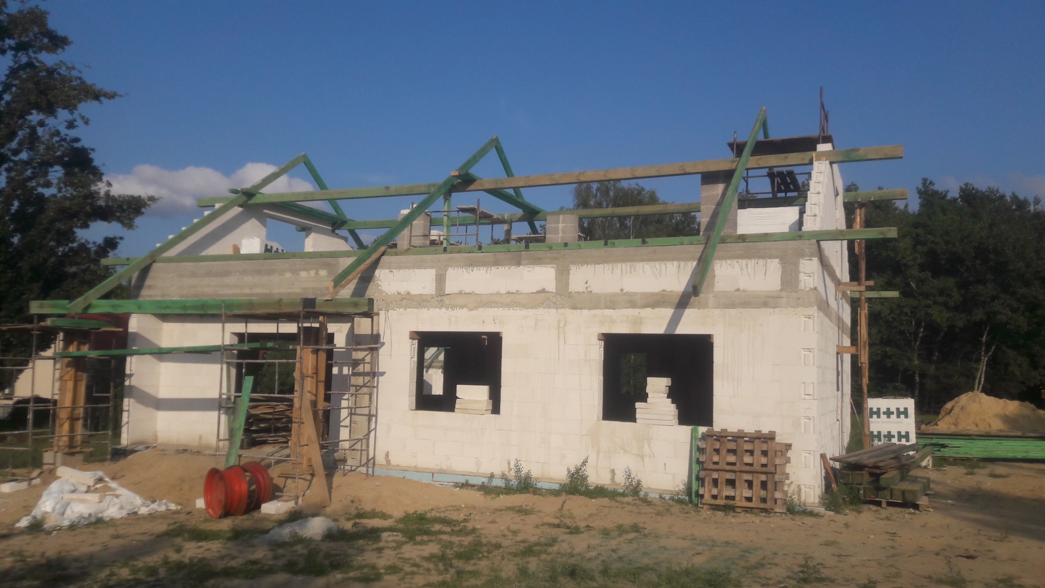 Surowy stan budynku jednorodzinnego z bloczków, z widoczną konstrukcją więźby dachowej z zielonych belek, niezadaszony, z otworami okiennymi i stertą bloczków w tle.