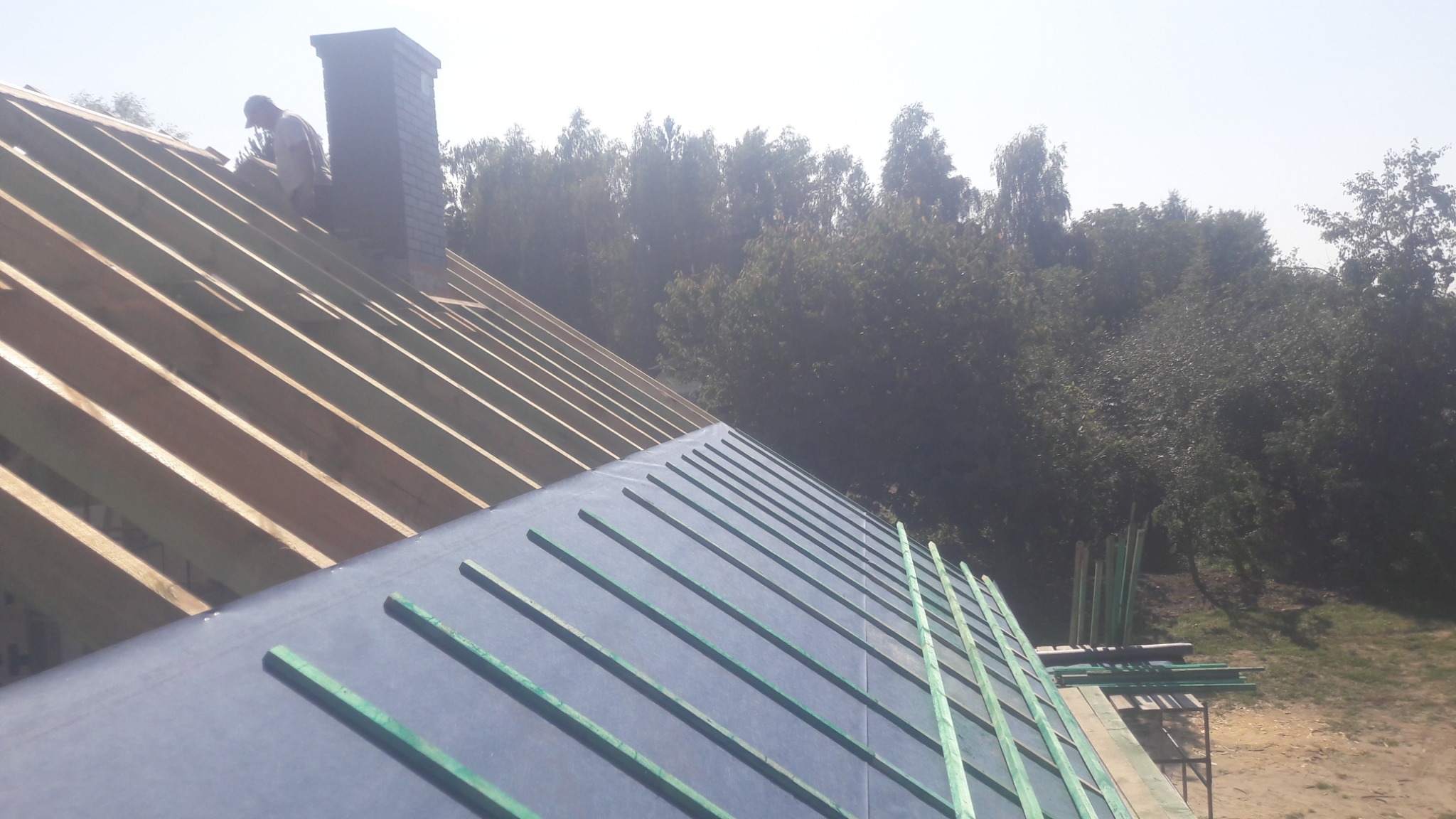 Montaż membrany dachowej z łatami na więźbie dachowej, w tle komin i drzewa.