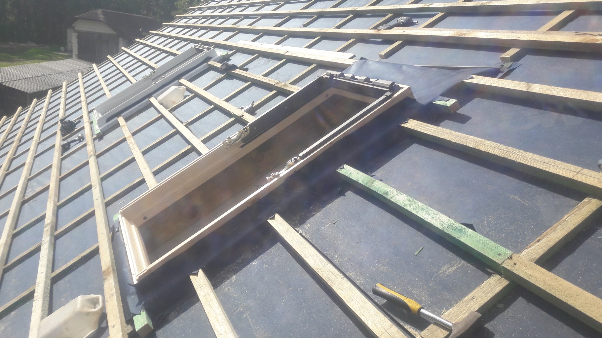 Dach w trakcie budowy z widocznymi łatami, membraną dachową i przygotowanymi otworami na okna dachowe. Widoczny młotek i poziomica.