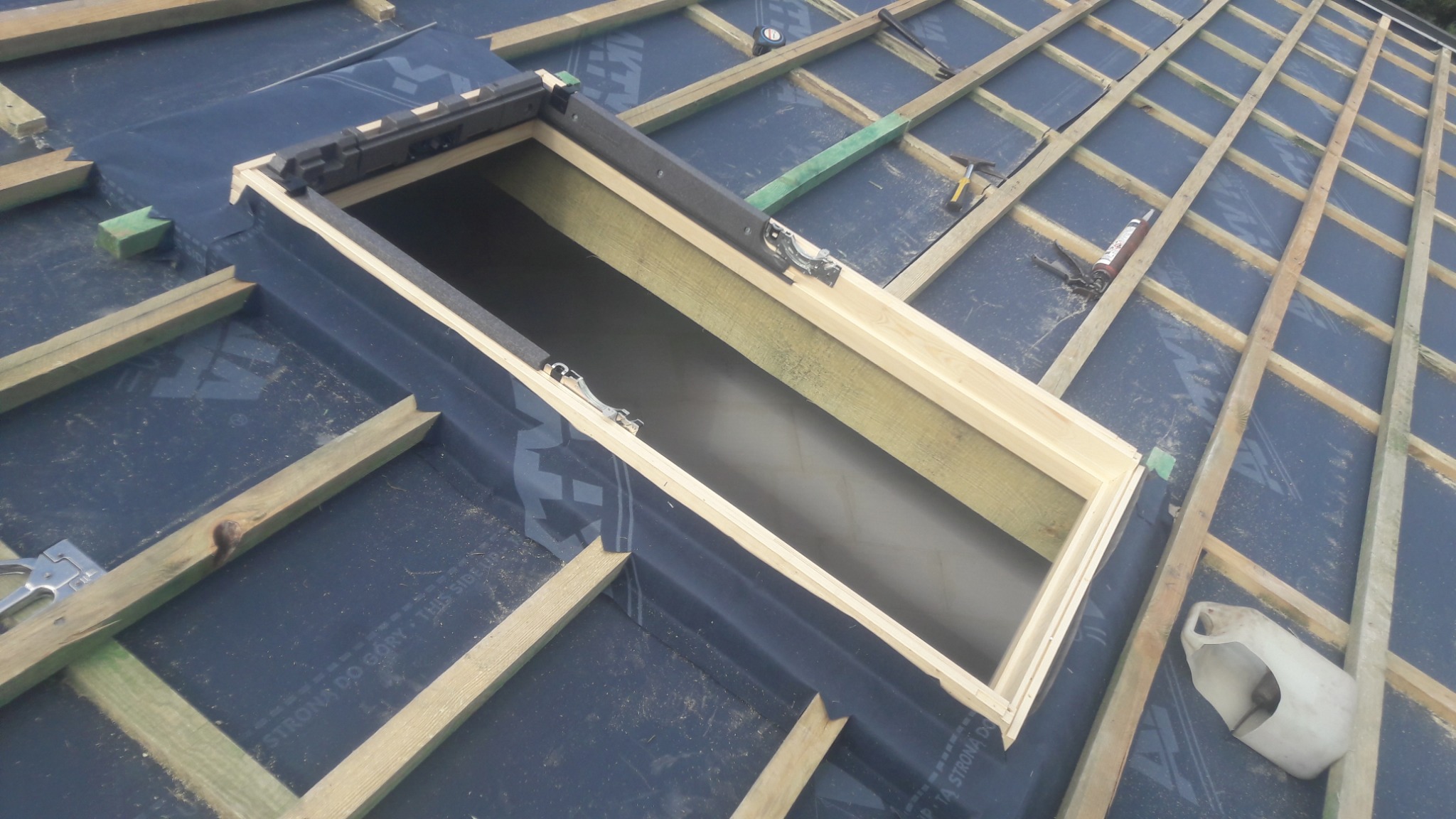 Montaż okna dachowego: widok na konstrukcję dachu z membraną, drewnianą ramą okienną i łatami. W tle widoczne narzędzia i akcesoria montażowe.