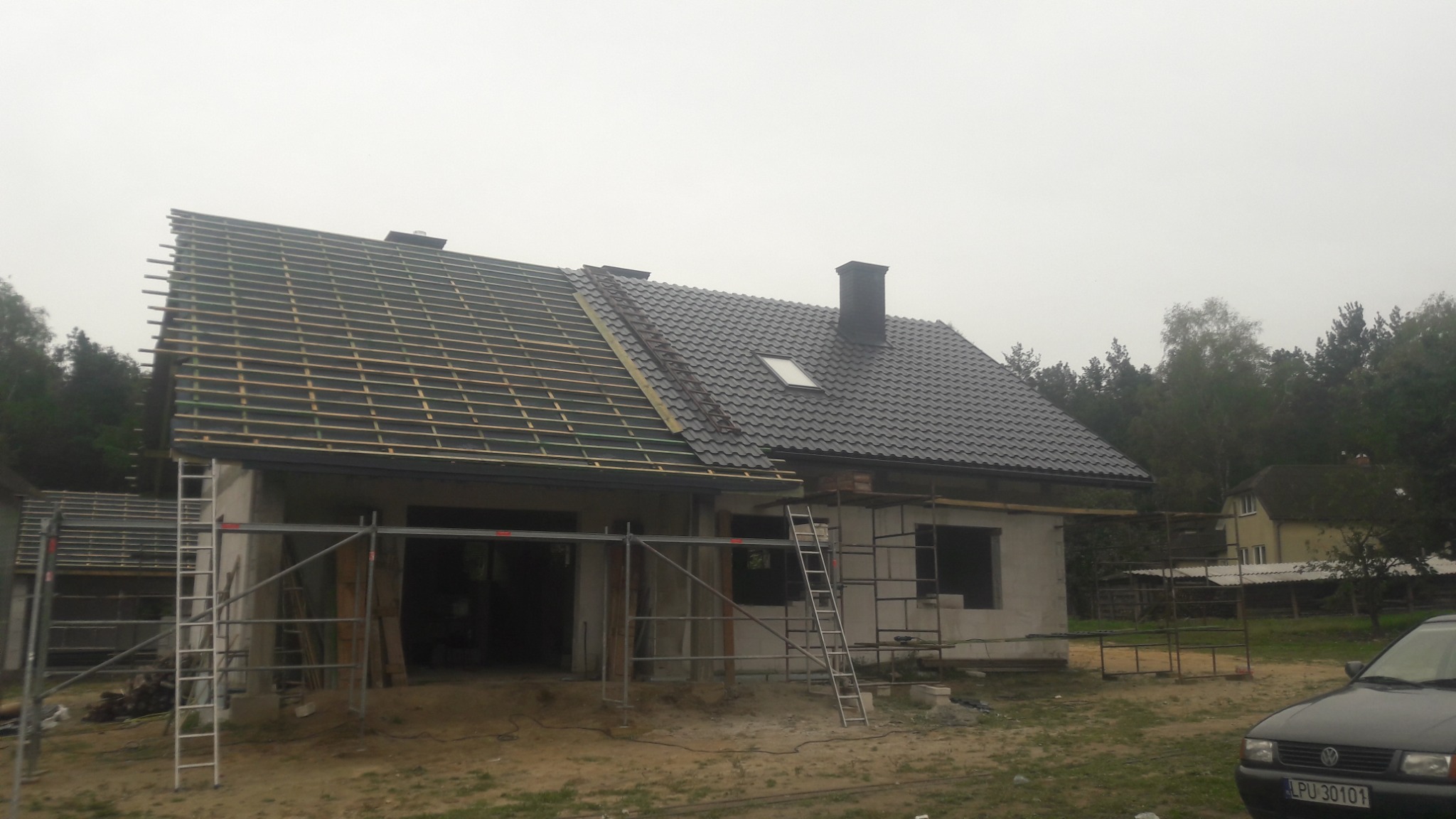 Dom w trakcie wymiany dachu, widoczna konstrukcja łat na jednej połaci i gotowe pokrycie dachowe z oknem na drugiej połaci, rusztowania wokół budynku, pochmurny dzień.