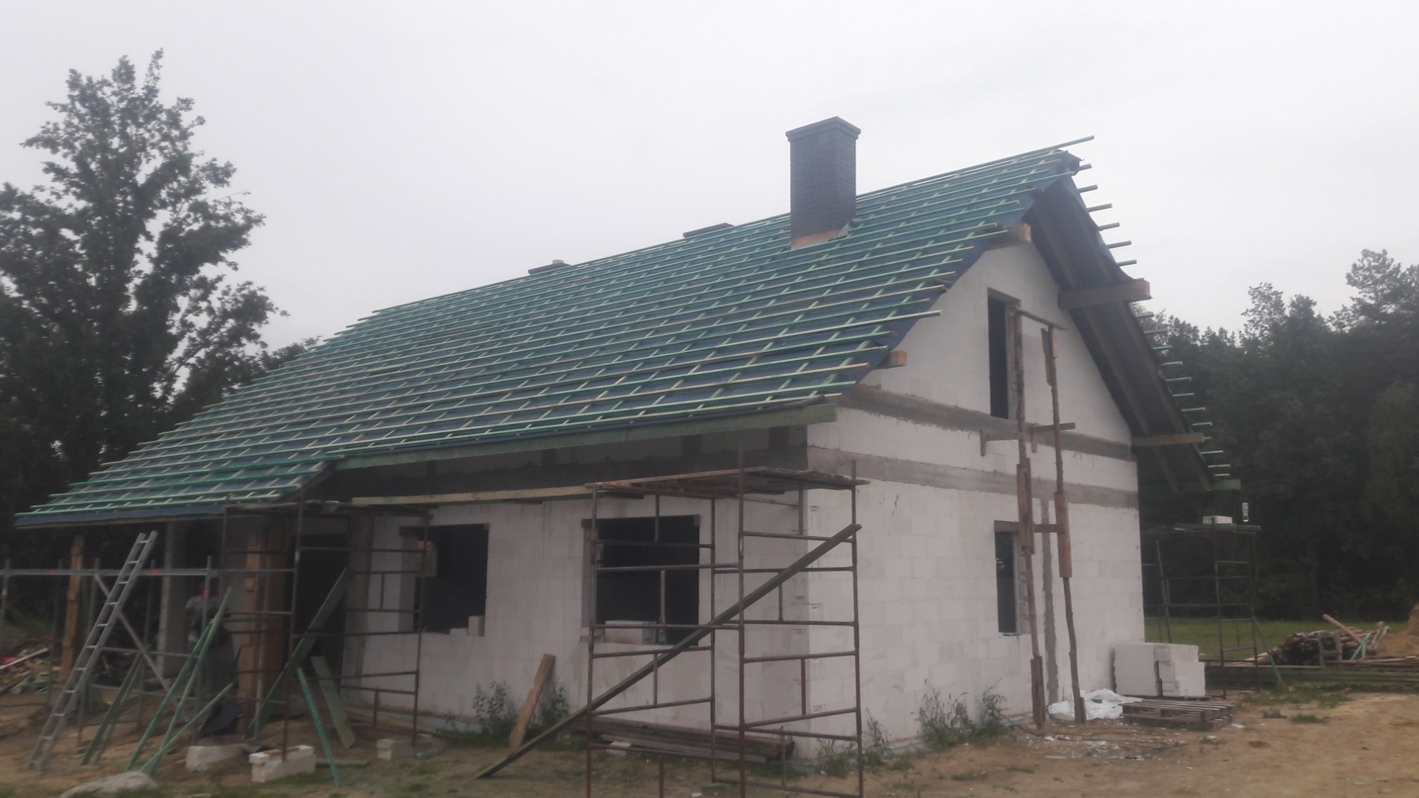 Dom w stanie surowym otwartym z widoczną konstrukcją dachu przygotowaną pod pokrycie, komin z cegły klinkierowej, ściany z betonu komórkowego, rusztowania.