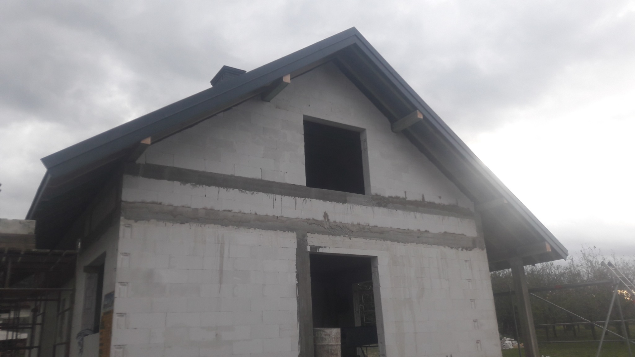 Surowy stan domu jednorodzinnego w trakcie budowy, widoczny dach z ciemnej blachy, ściany z białych bloczków, niezadaszony otwór okienny na poddaszu, szare niebo w tle.