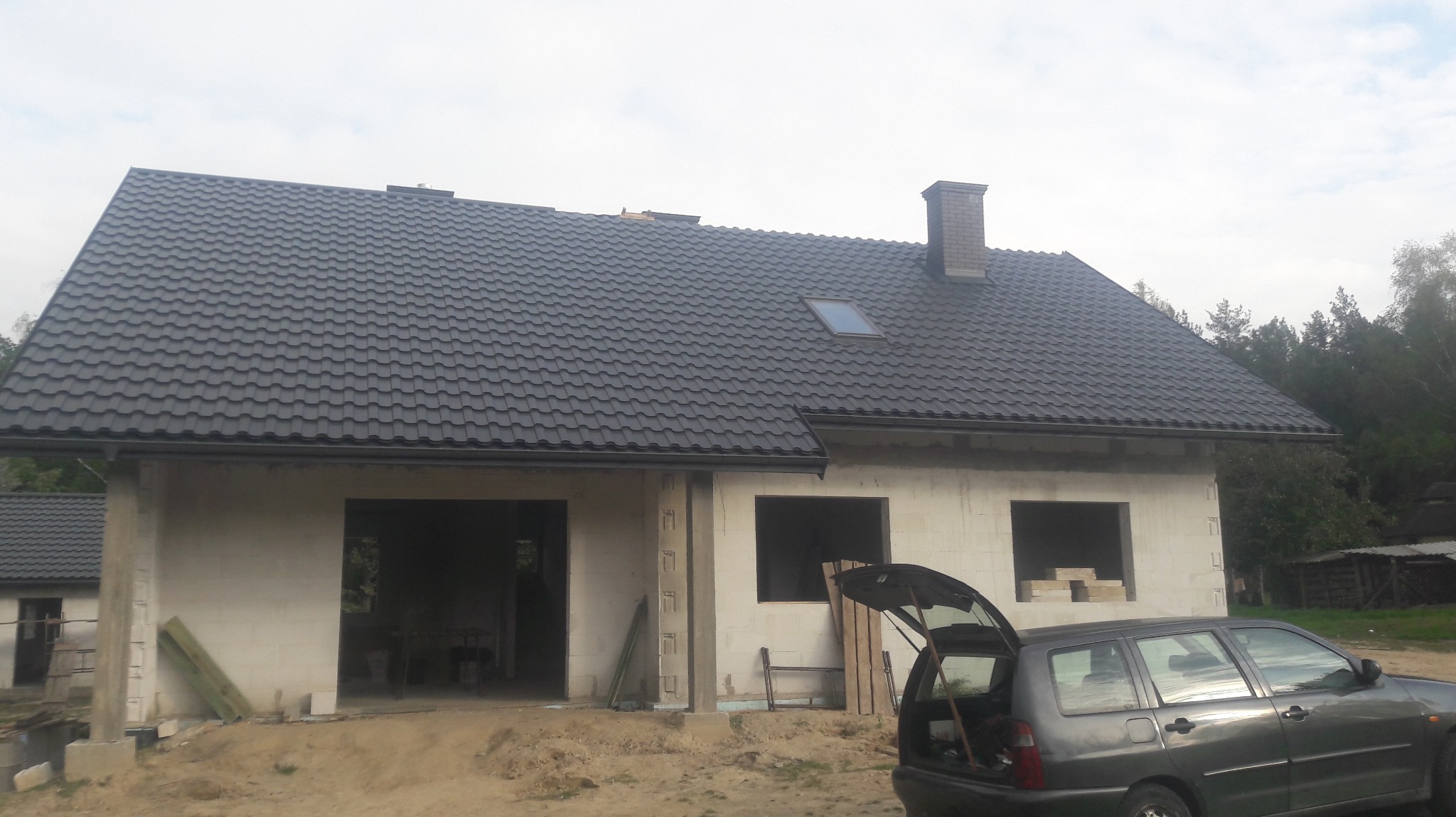Nowy, grafitowy dach z oknem połaciowym na surowym domu jednorodzinnym w trakcie budowy, widok z boku z otwartą klapą bagażnika samochodu osobowego.