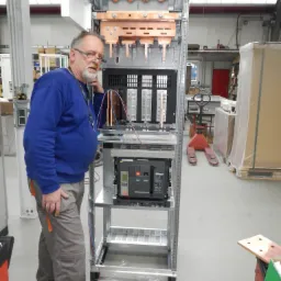 Elektryk w okularach, ubrany w niebieską bluzę i szare spodnie robocze, stoi obok rozdzielnicy elektrycznej w trakcie montażu. Widoczne są miedziane szyny i elementy zabezpieczające. W tle hala...