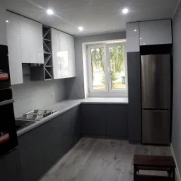 Nowoczesna, szaro-biała kuchnia z zabudową, z widoczną płytą grzewczą, piekarnikiem i lodówką. Na podłodze panele imitujące drewno. W oknie widok na zieleń.