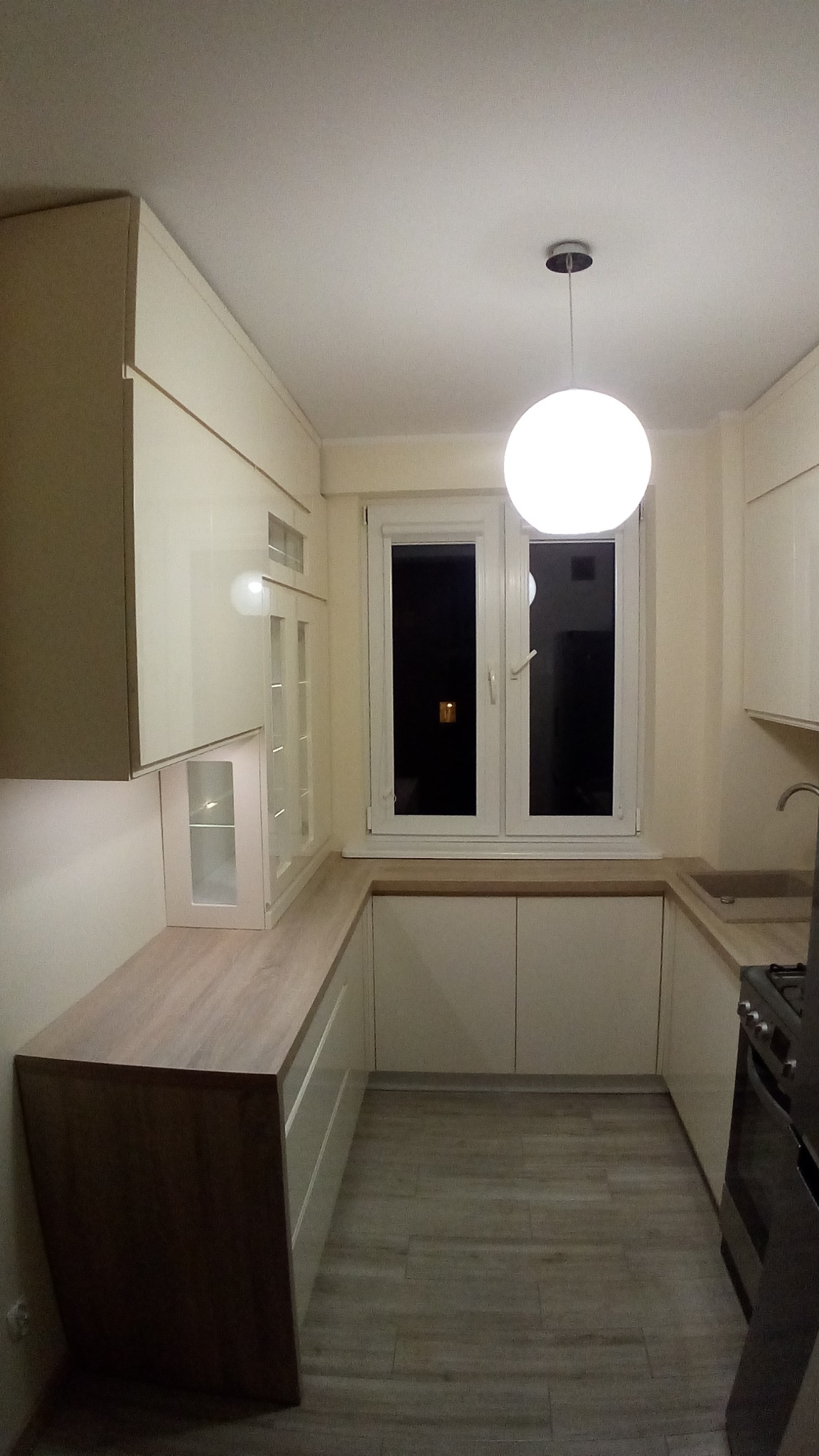 Ustawna kuchnia w bloku z jasnymi, lakierowanymi frontami, drewnianym blatem pod oknem i wiszącą, okrągłą lampą.