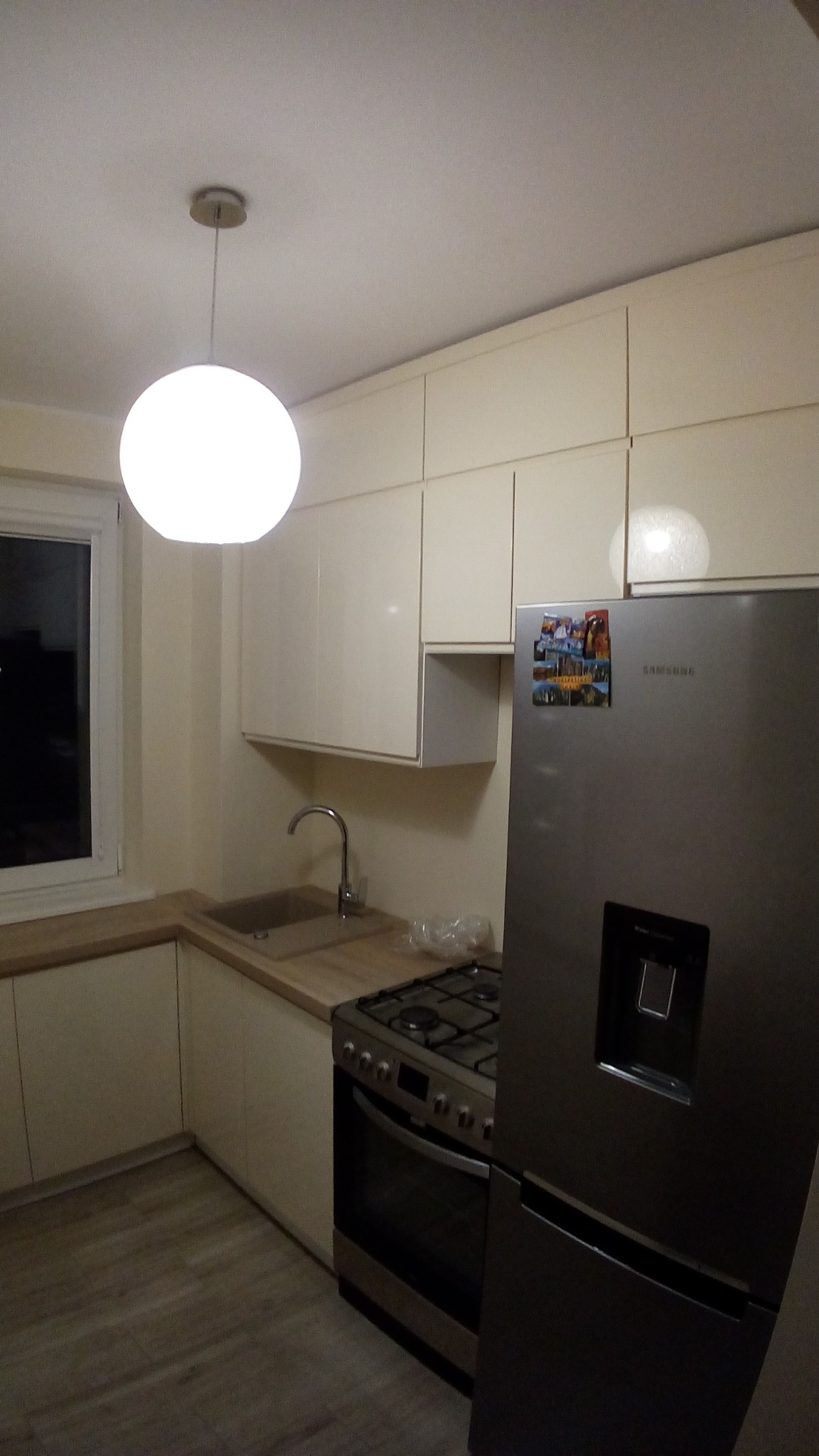 Jasna kuchnia z beżowymi szafkami i drewnianym blatem, widoczny zlew, płyta gazowa i lodówka Samsung z magnesami na drzwiach.