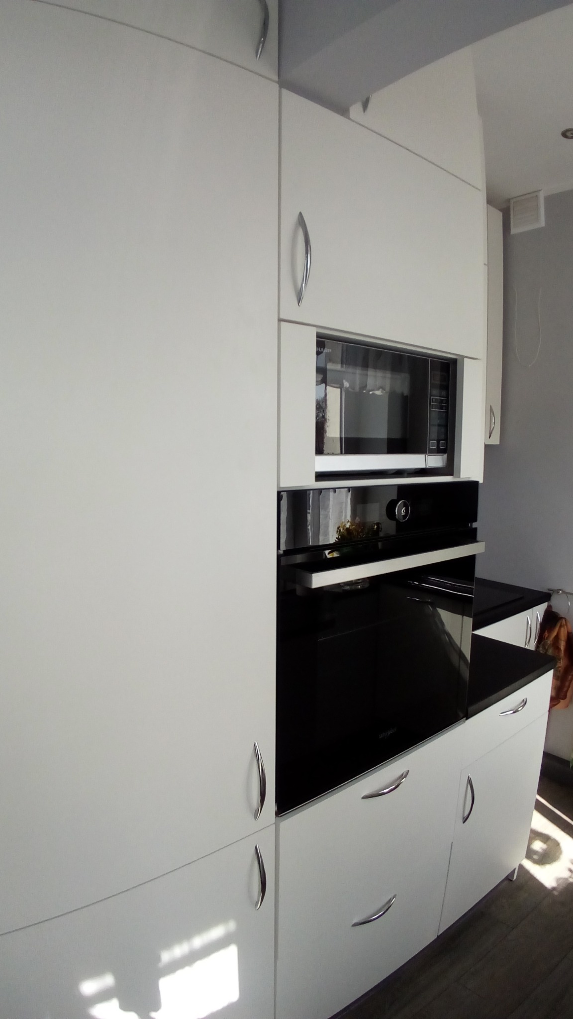 Białe, lakierowane szafki kuchenne z mikrofalówką i piekarnikiem w zabudowie, minimalistyczne uchwyty, czarny blat.