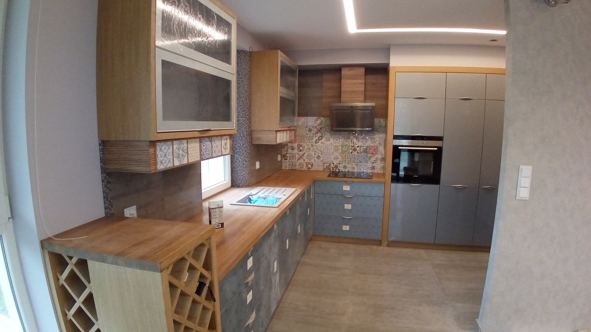 Nowoczesna zabudowa kuchenna z drewnianym blatem, szafkami górnymi z folią ochronną i dekoracyjnymi płytkami pod oknem, uzupełniona o regał na wino.