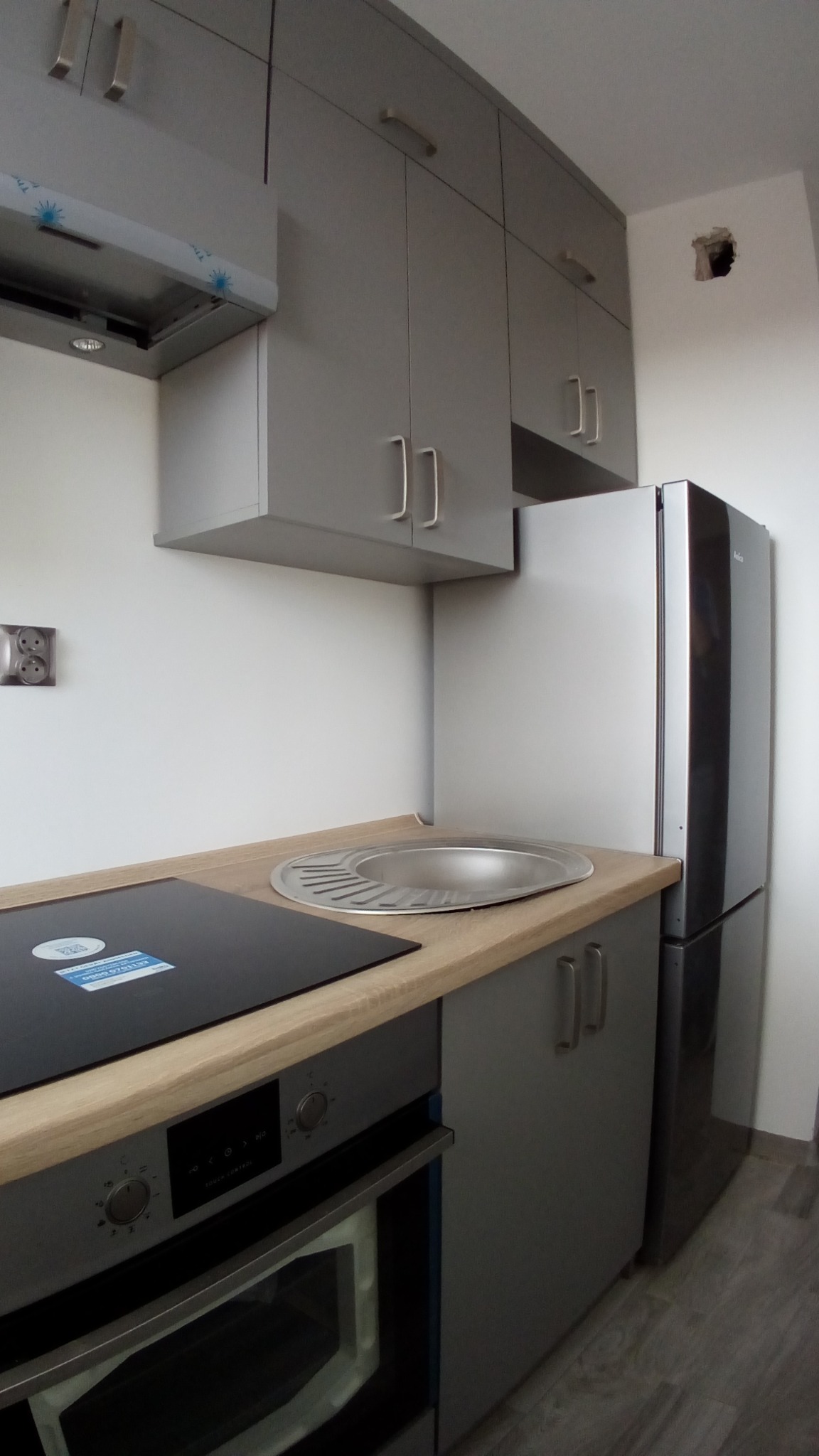 Szara zabudowa kuchenna z płytą indukcyjną, zlewem i piekarnikiem, widoczny fragment lodówki i górne szafki, niedokończone wykończenie ściany.