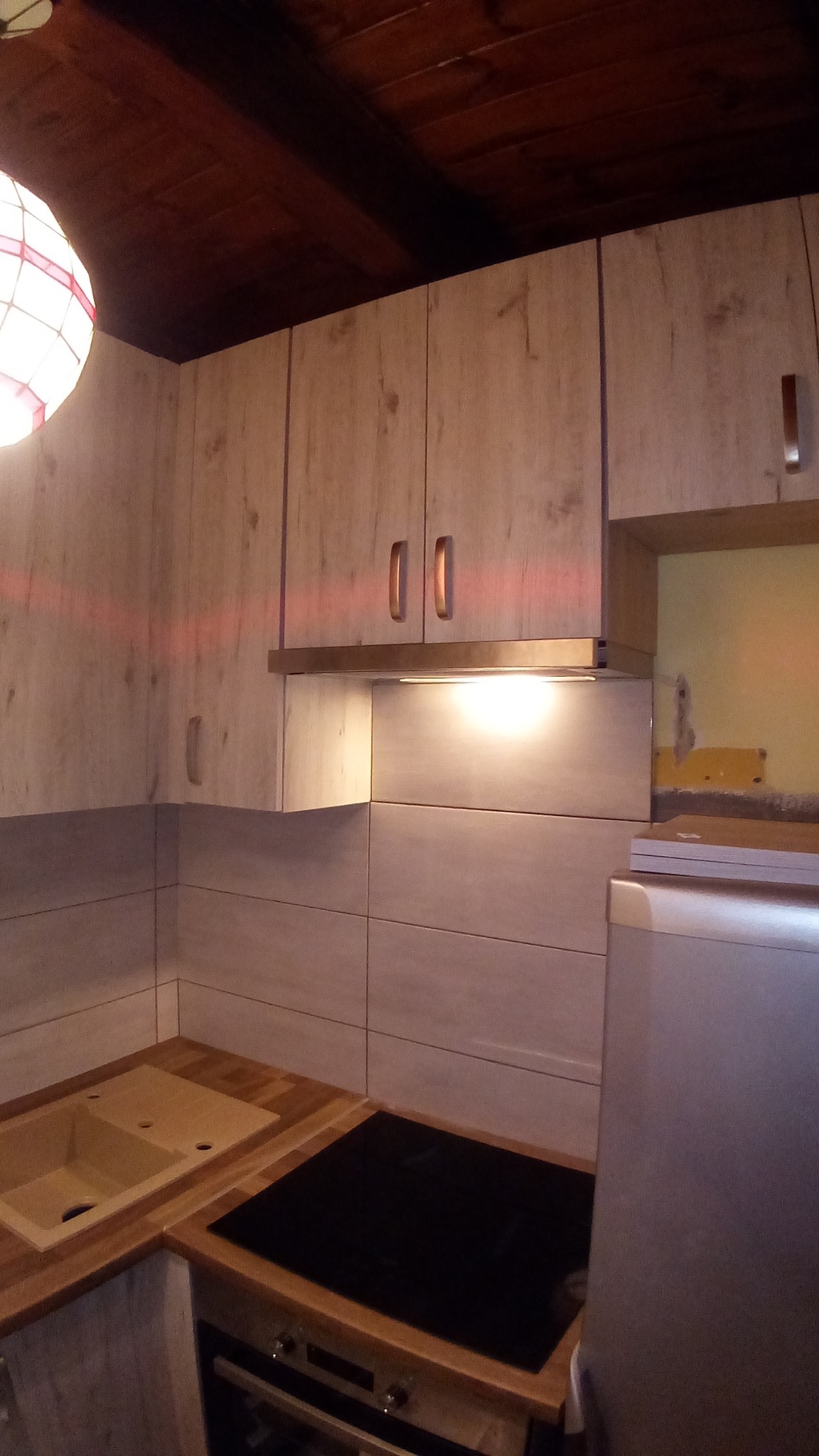 Kuchenna zabudowa z drewnopodobnymi szafkami, ceramicznym zlewem i płytą indukcyjną, widoczne fragmenty instalacji elektrycznej.