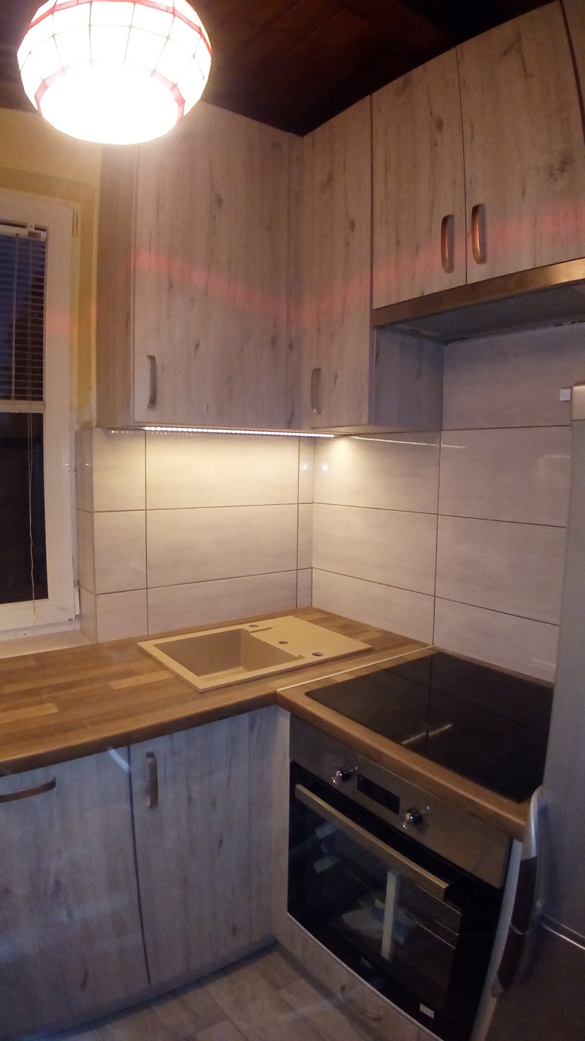 Fragment zabudowy kuchennej z drewnopodobnymi frontami, zlewem z ociekaczem i płytą grzewczą, oświetlony listwą LED pod szafkami wiszącymi.