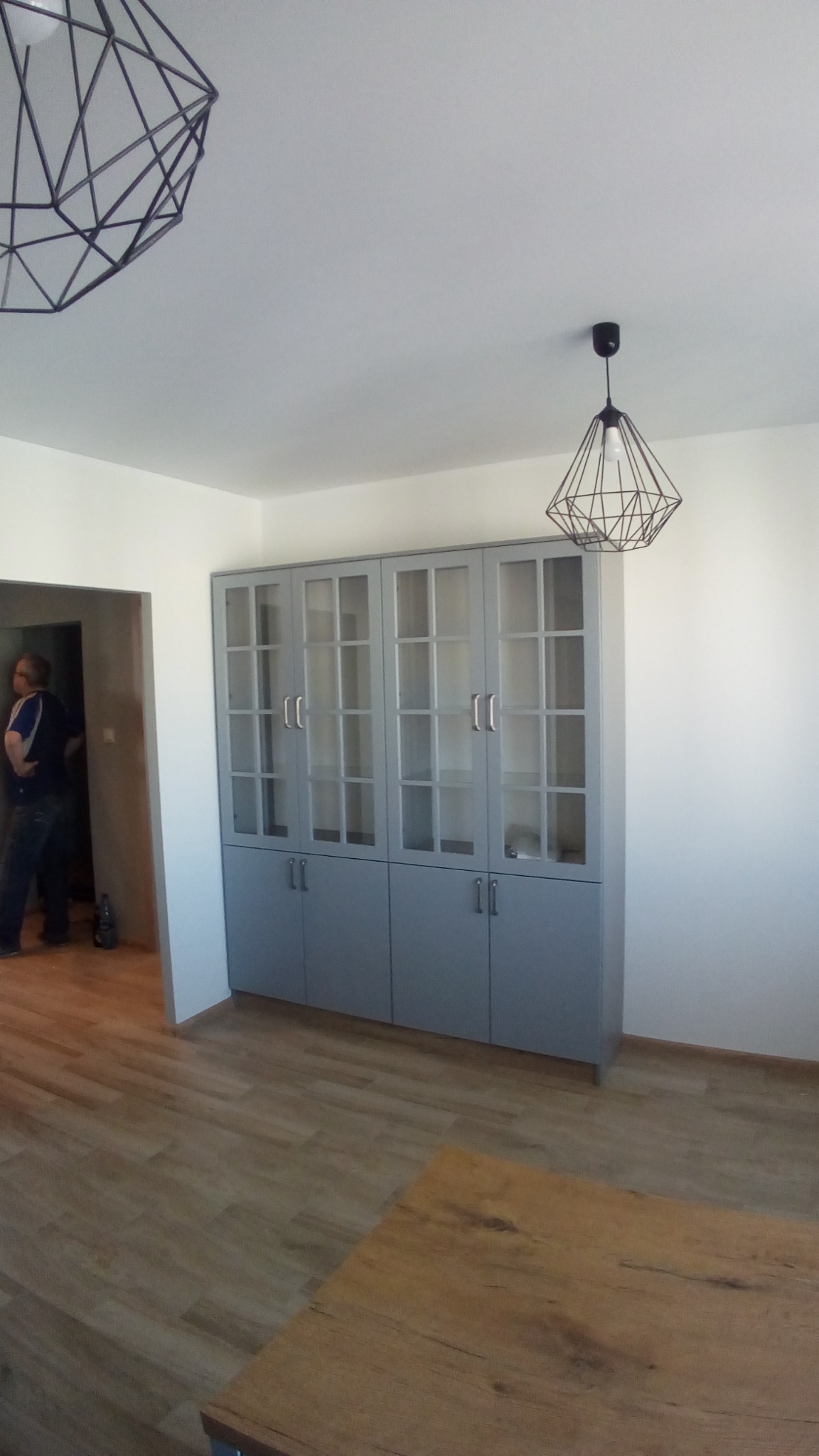 Szara szafa z przeszklonymi drzwiami, podzielona na segmenty, z drewnianym blatem stołu w dolnej części kadru i geometrycznymi lampami wiszącymi na suficie, widok na osobę w przejściu.