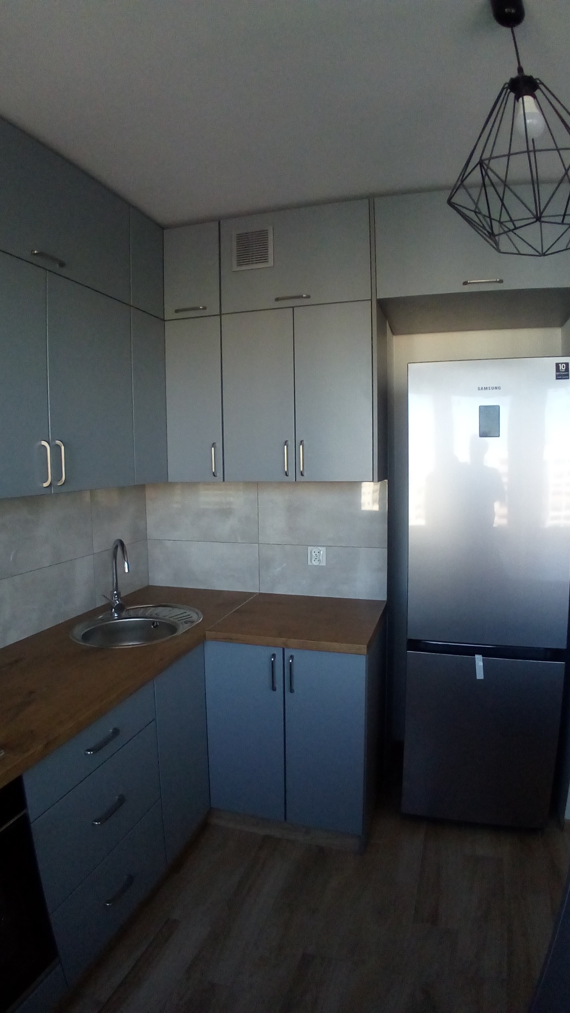 Szara kuchnia na wymiar z drewnianym blatem, chromowanym zlewem i nowoczesną lampą wiszącą, uzupełniona o srebrną lodówkę Samsung.