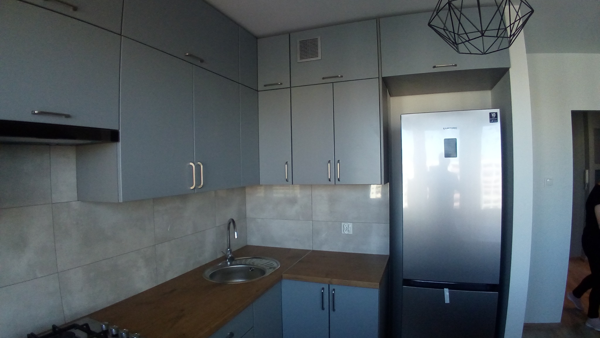 Szara kuchnia z drewnianym blatem i srebrną lodówką Samsung, oświetlona nowoczesną lampą sufitową z czarnych drutów.