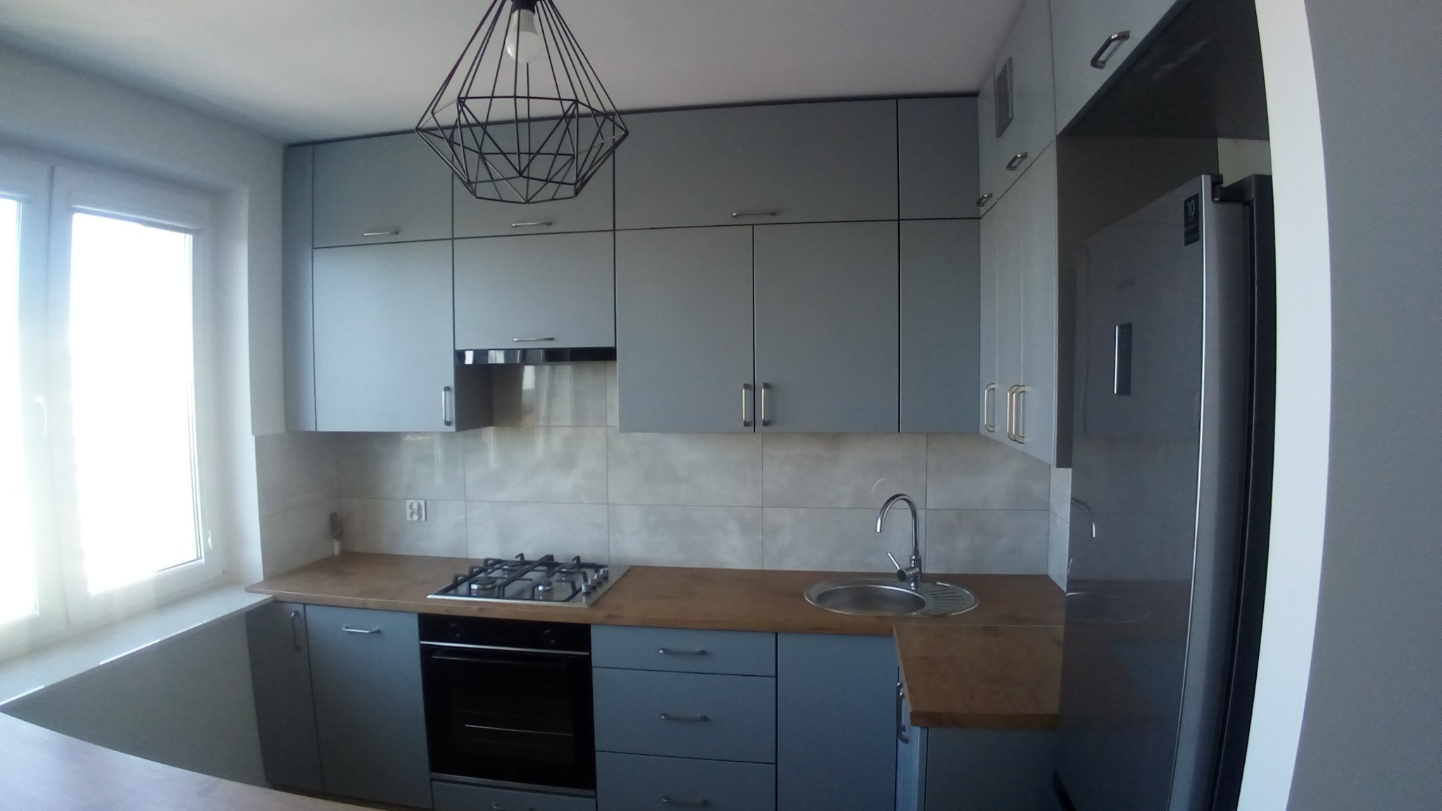 Nowoczesna kuchnia w szarościach: zabudowa z górnymi szafkami pod sufit, drewniany blat, płyta gazowa, zlewozmywak i wisząca lampa w geometrycznym stylu.