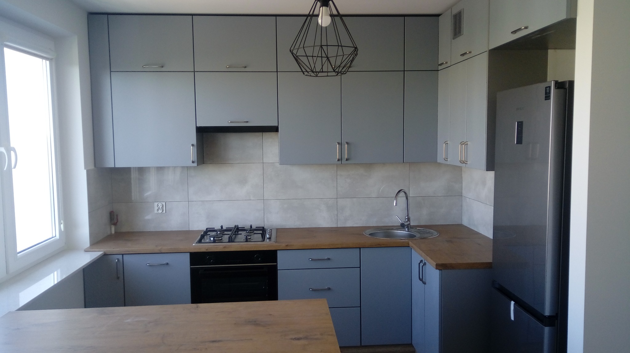 Jasnoszara kuchnia z drewnianymi blatami, nowoczesna lampa wisząca, widoczny fragment okna i lodówka.