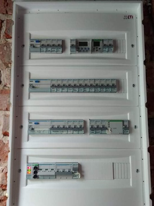Rozdzielnica elektryczna w białej obudowie, zamontowana na tle ściany z czerwonej cegły, widoczne zabezpieczenia i liczniki, niektóre z włączonymi diodami.