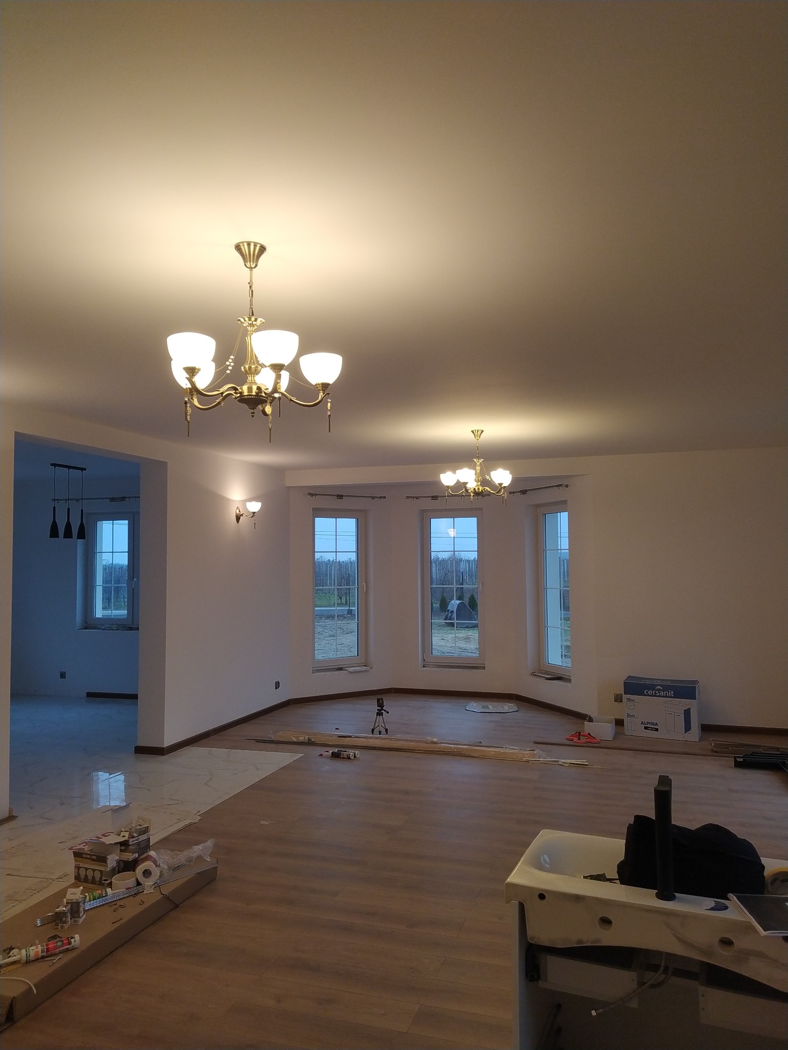 Wnętrze domu w trakcie wykańczania: położona częściowo podłoga, widoczne narzędzia i materiały budowlane, zamontowane dwa żyrandole w stylu retro oraz kinkiet ścienny, okna z widokiem na ogród.