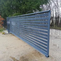 Przesuwna, pozioma brama wjazdowa z szarych, metalowych profili, widoczny mechanizm zębatkowy, tło stanowią drzewa i mokra nawierzchnia.