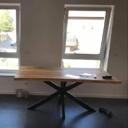 Stół z drewnianym blatem i metalową podstawą w kształcie gwiazdy, w tle okna i narzędzia na podłodze.