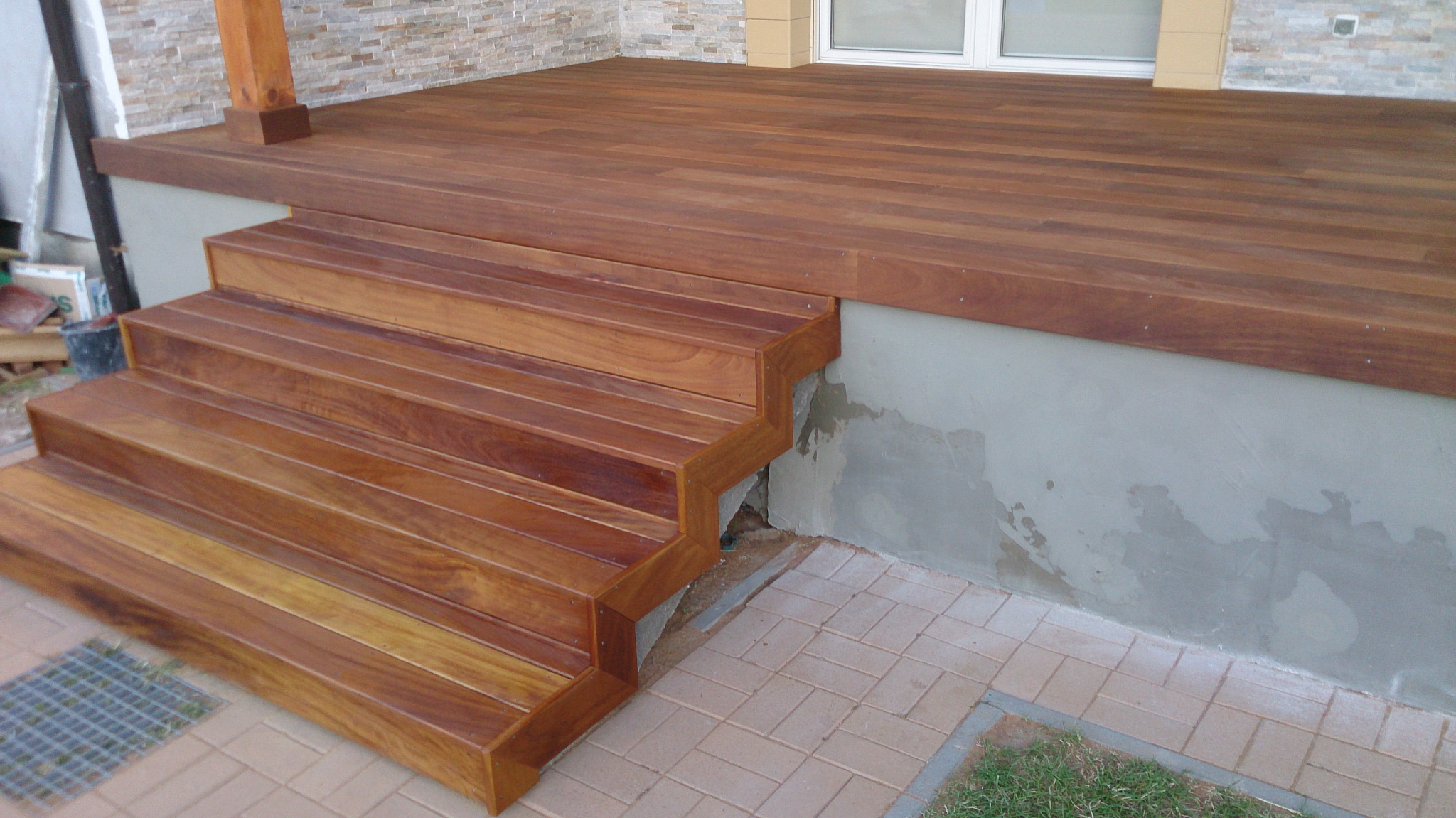 Taras z deski IROKO z drewnianymi schodami wejściowymi, widok z boku na szary fundament i kostkę brukową