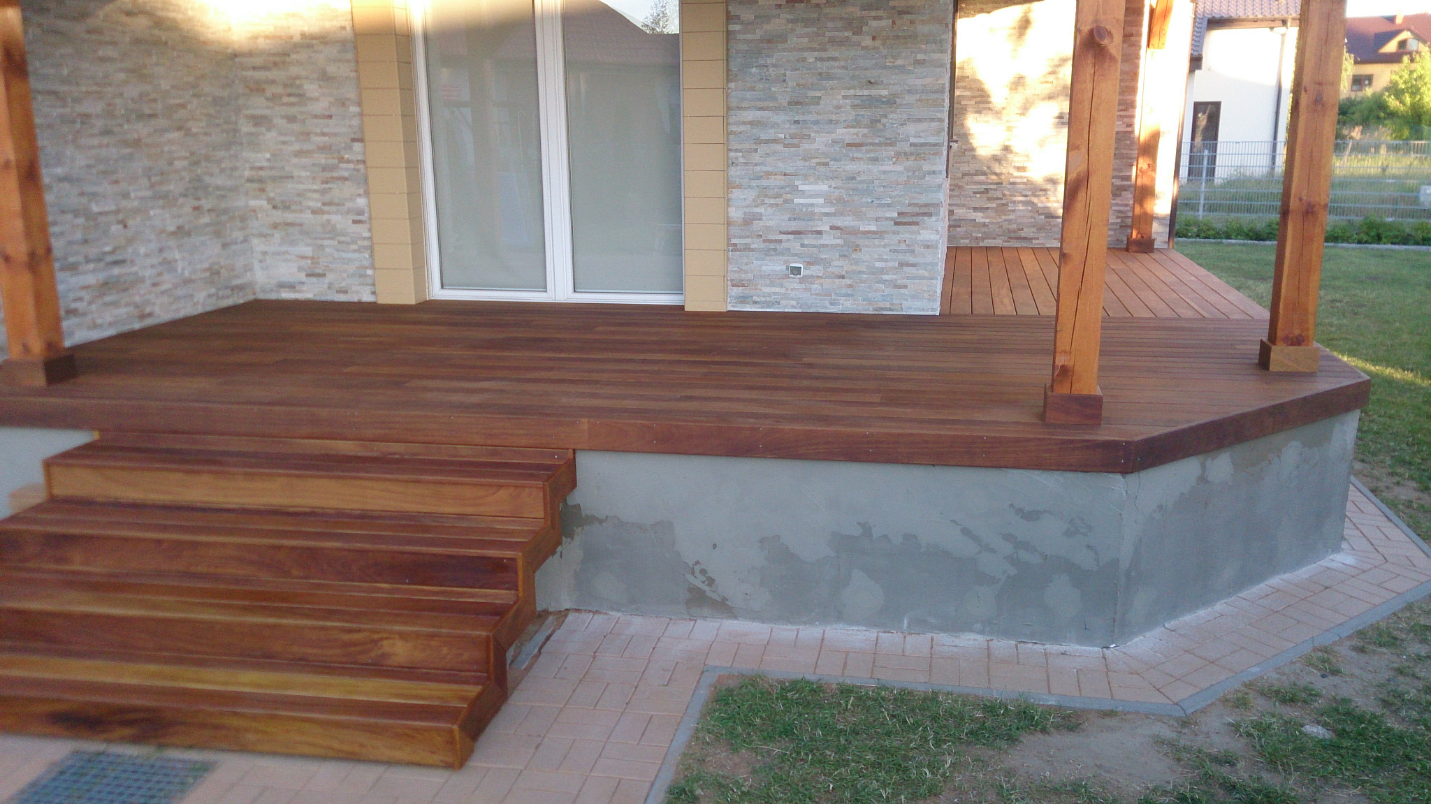 Wykończony taras z deski IROKO z drewnianymi schodami i filarami, widoczne jasne płytki elewacyjne na ścianie budynku i trawnik w tle.