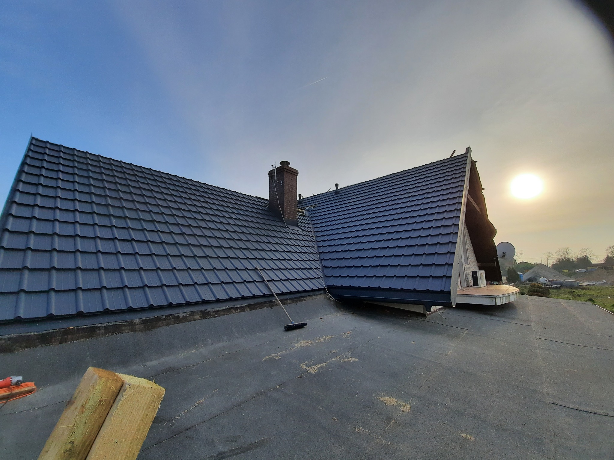 Nowy, grafitowy dach z blachodachówki modułowej, widok z dachu sąsiedniego, w tle błękitne niebo z promieniami zachodzącego słońca, na pierwszym planie deski i szczotka dekarska.