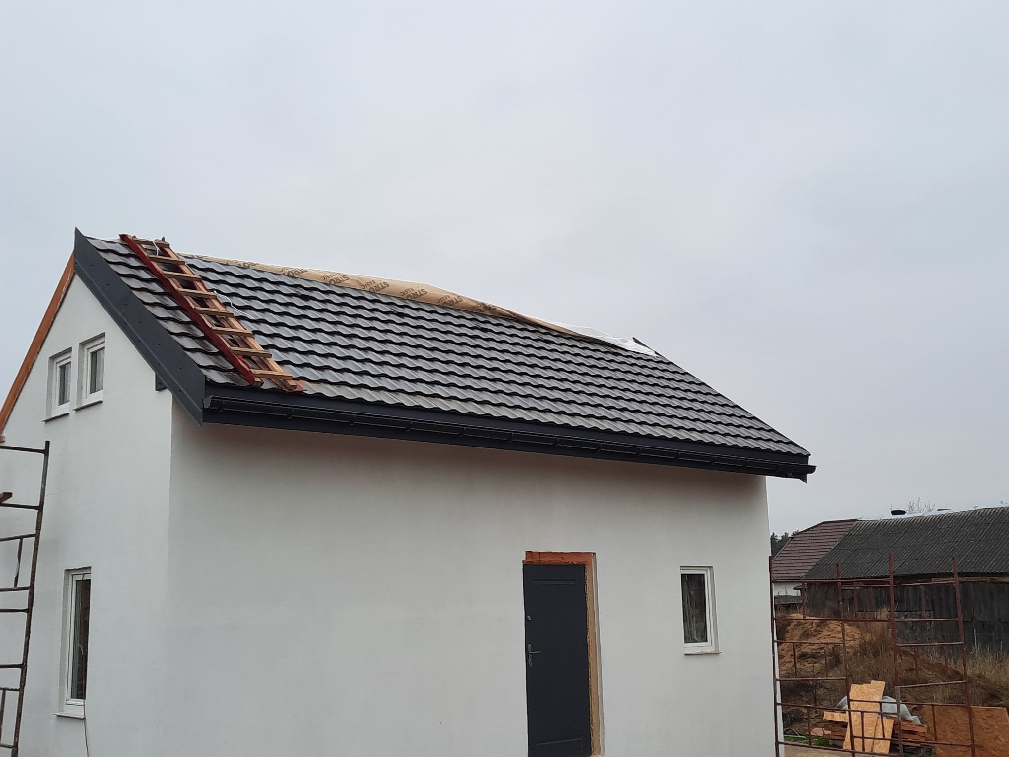 Nowy, grafitowy dach z blachodachówki na białym budynku gospodarczym, częściowo przykryty folią ochronną, z drewnianą drabiną opartą o krawędź dachu. Widoczny fragment rynny i drabina przystawna...