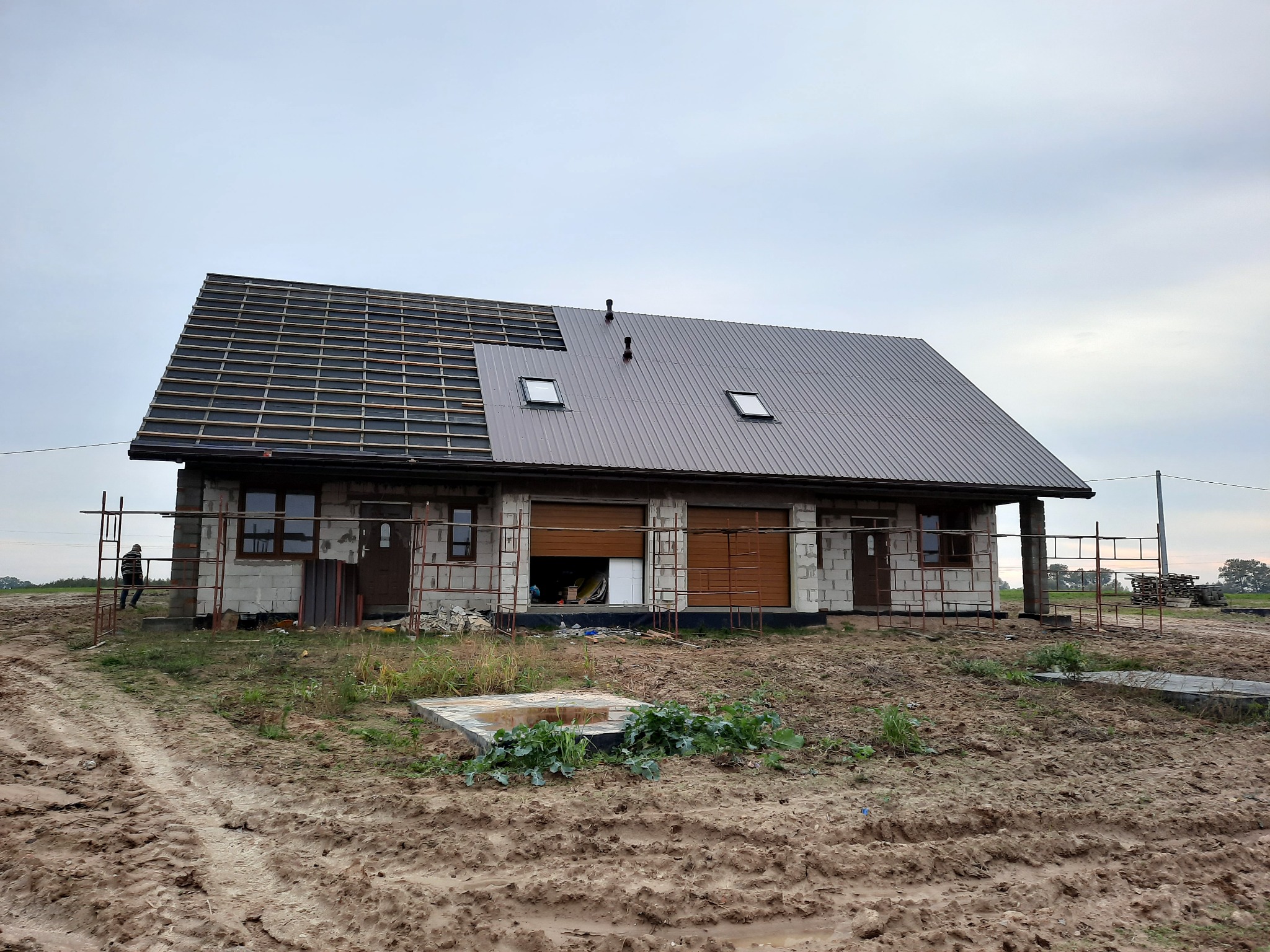 Dom w trakcie budowy z częściowo położonym brązowym dachem z blachy trapezowej i oknami dachowymi; widoczne rusztowania i teren wokół budynku w stanie surowym.